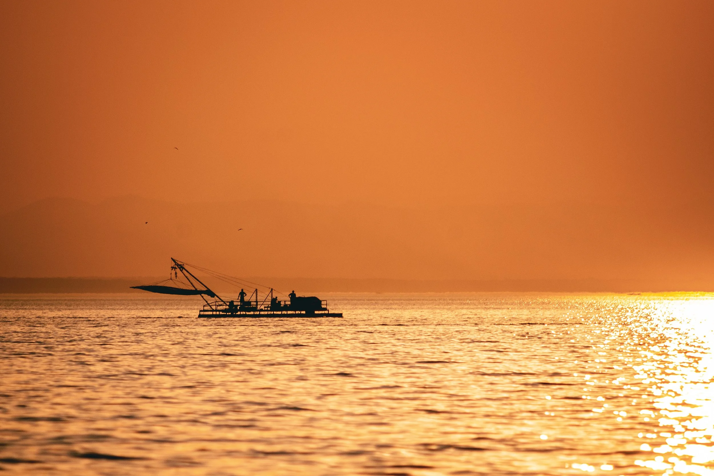 Silhouette of a fishing boat on calm water during sunset with orange sky.