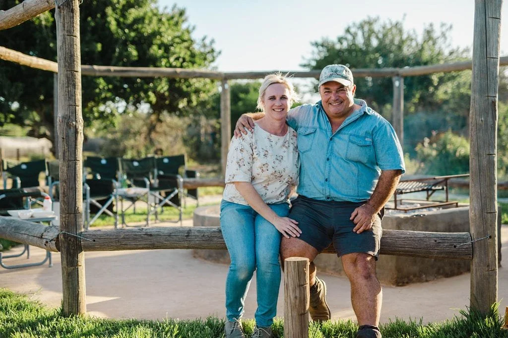 A smiling couple sitting on a wooden bench in a park or backyard, with trees and outdoor seating in the background, during daylight.