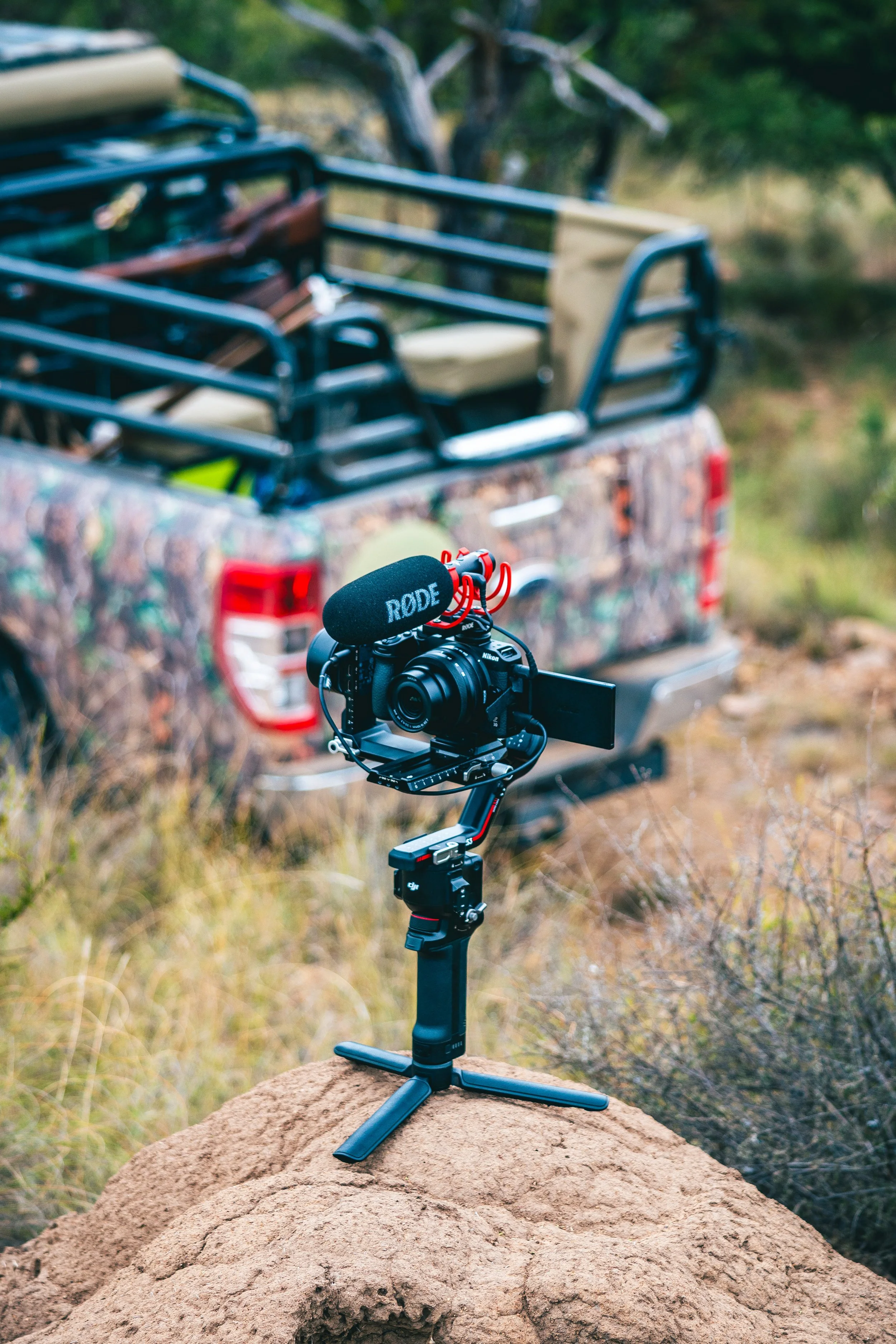 Camera mounted on a small tripod with a shoulder rig, placed on a rock, filming a camouflage truck with a cage on the bed, in a natural outdoor setting.