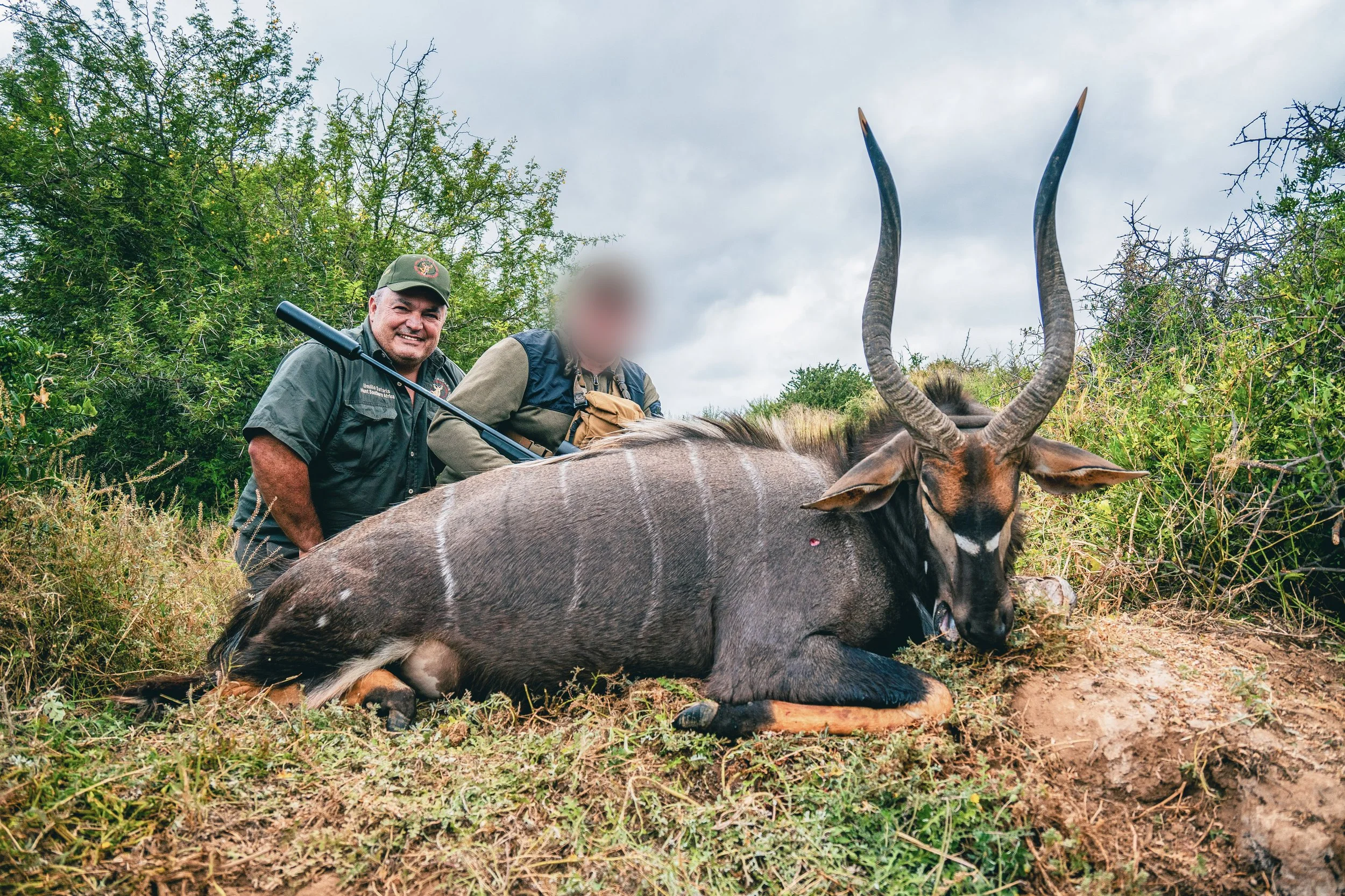 Two men stand behind a dead antelope with long, curved horns, lying on the ground in tall grass and shrubs in a wilderness area.
