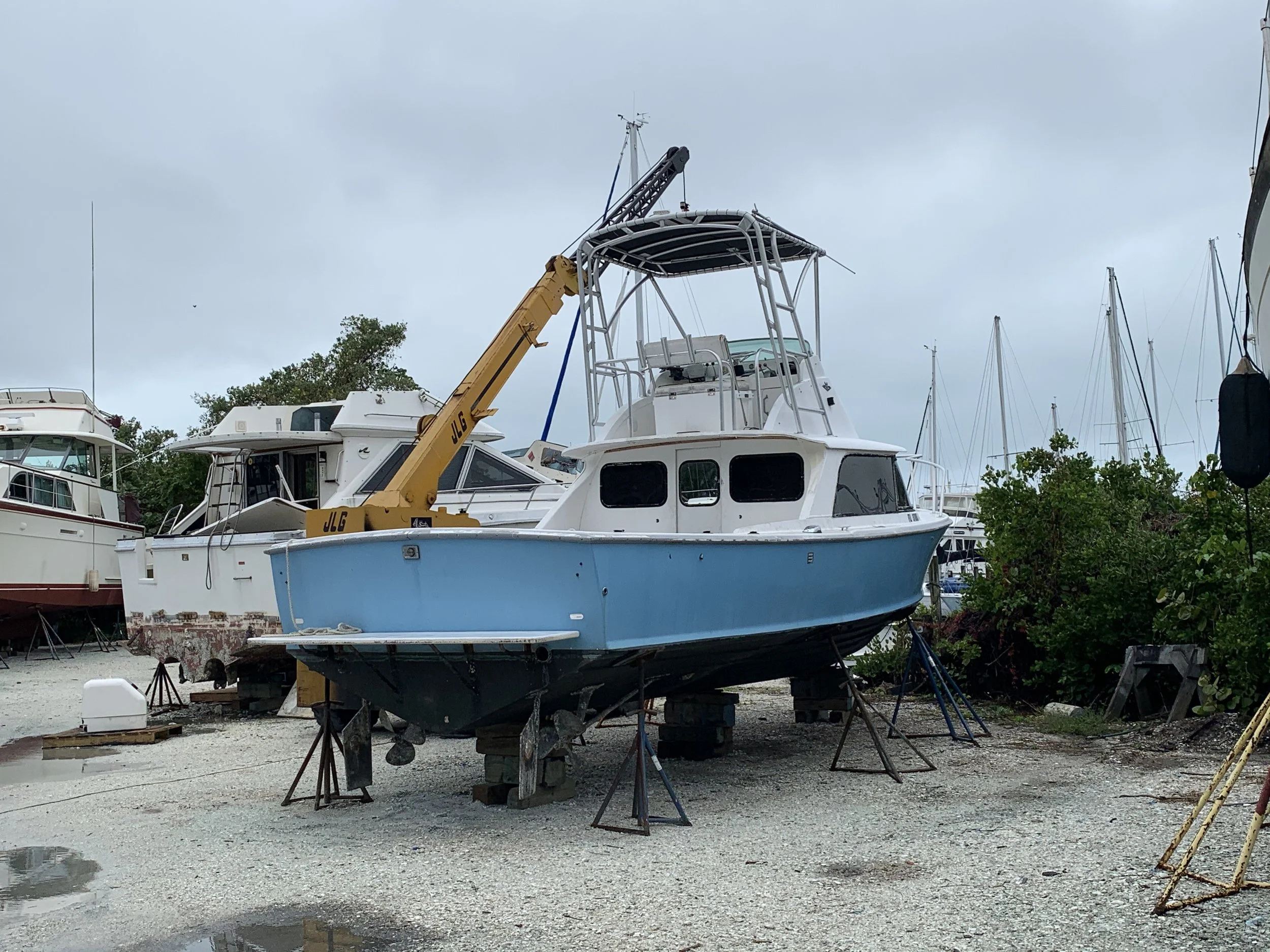 A boat is on land in a boatyard, lifted on supports, with a yellow crane nearby. Other boats are visible in the background, and the sky is overcast.