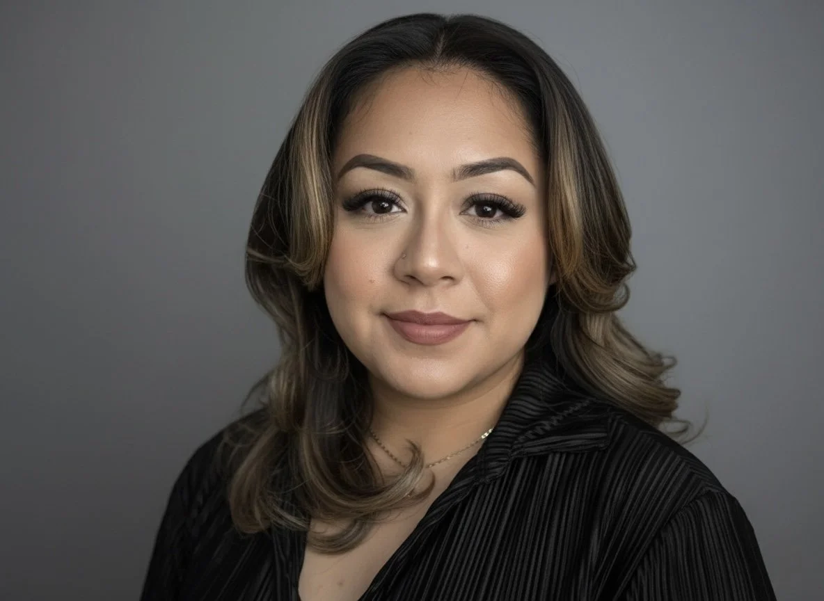 A woman with shoulder-length brown hair, makeup, and a black top, looking at the camera against a gray background.