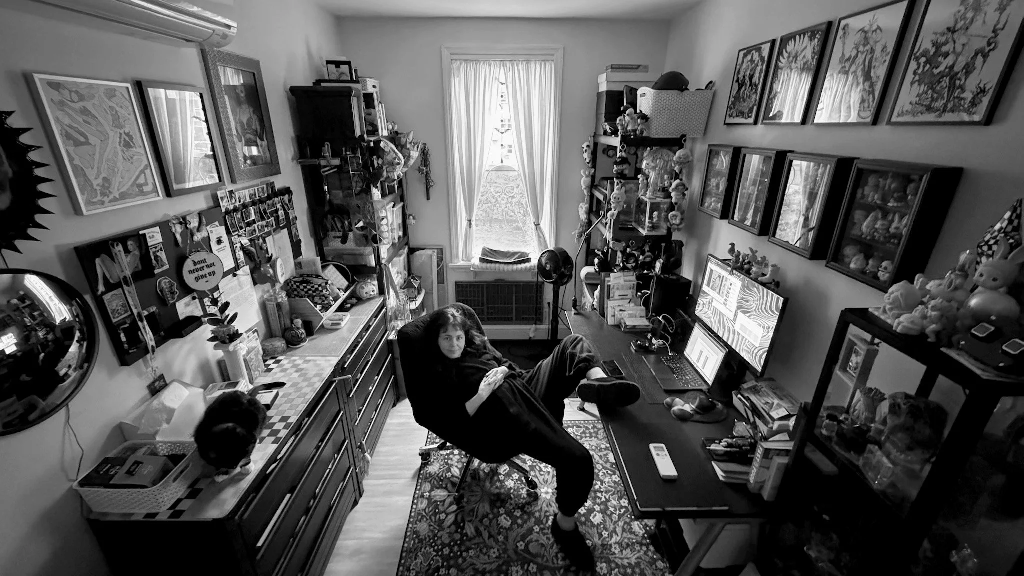 A woman with dark hair sitting in a computer chair in a cluttered home office, looking at the camera, with two monitors, a laptop, and various decorative items and toys around the room.
