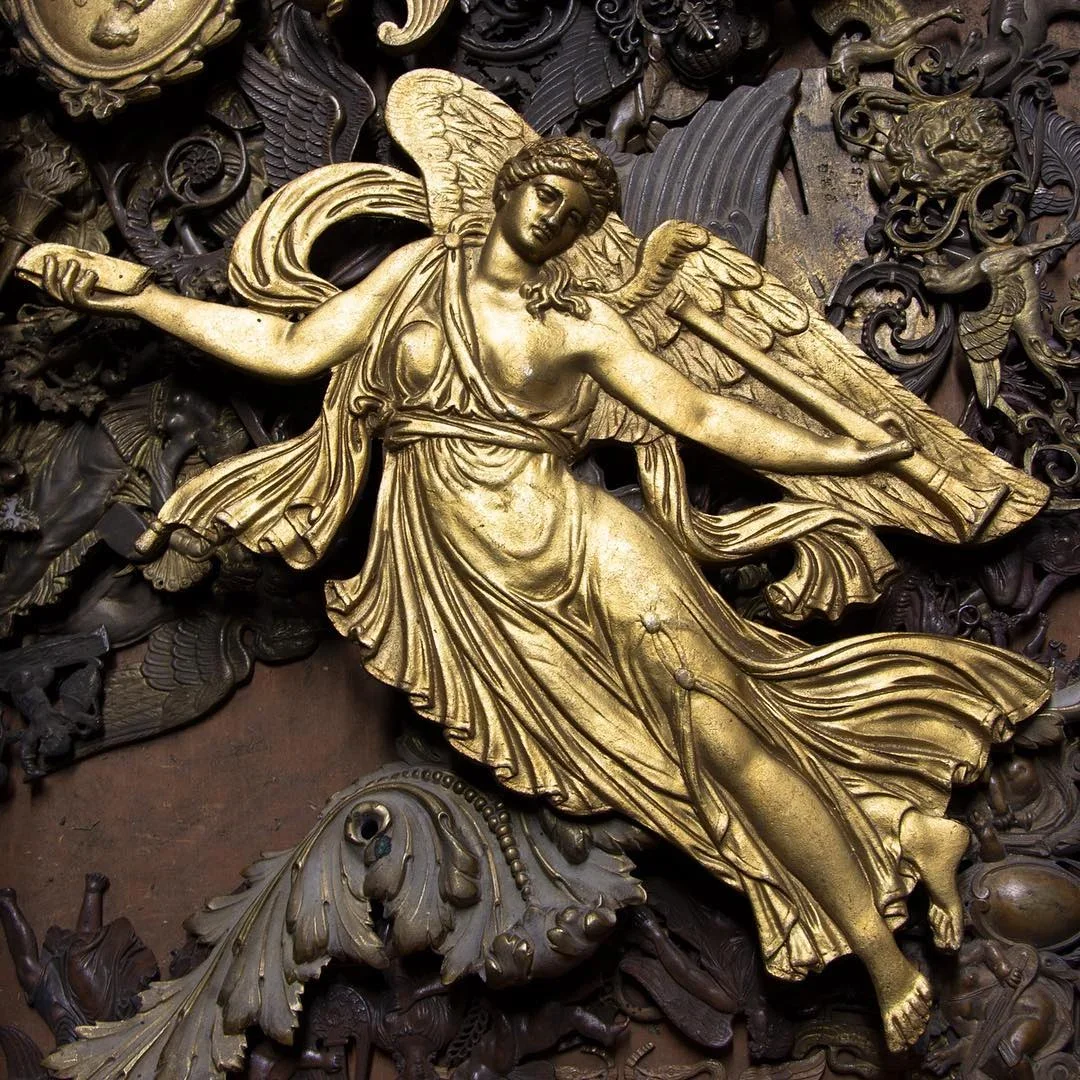A detailed, ornate sculpture of a winged female figure in gold, surrounded by intricate, dark relief carvings of various mythical and fantastical creatures.