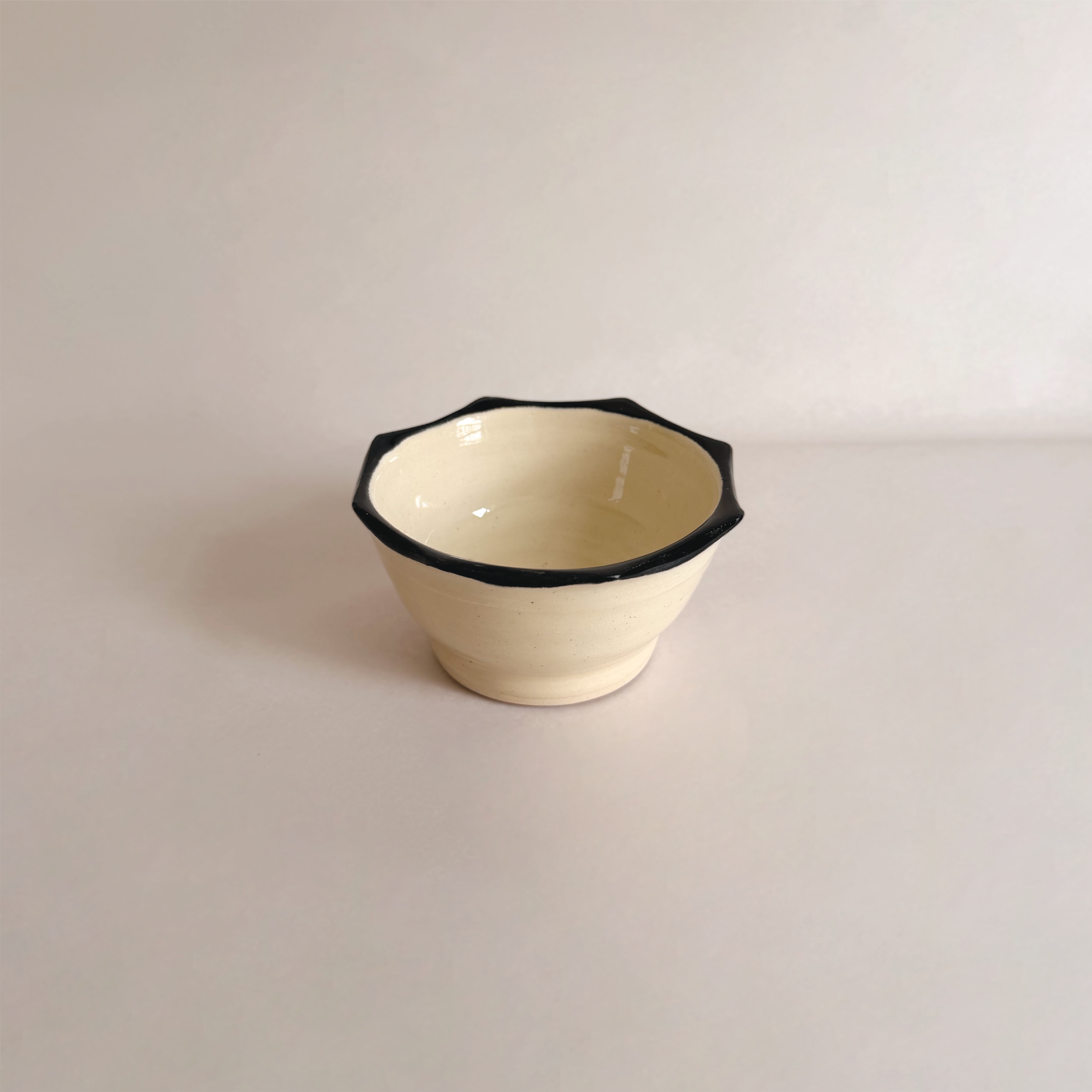 A small ceramic bowl with a black rim, cream-colored, and empty, placed on a plain white surface.