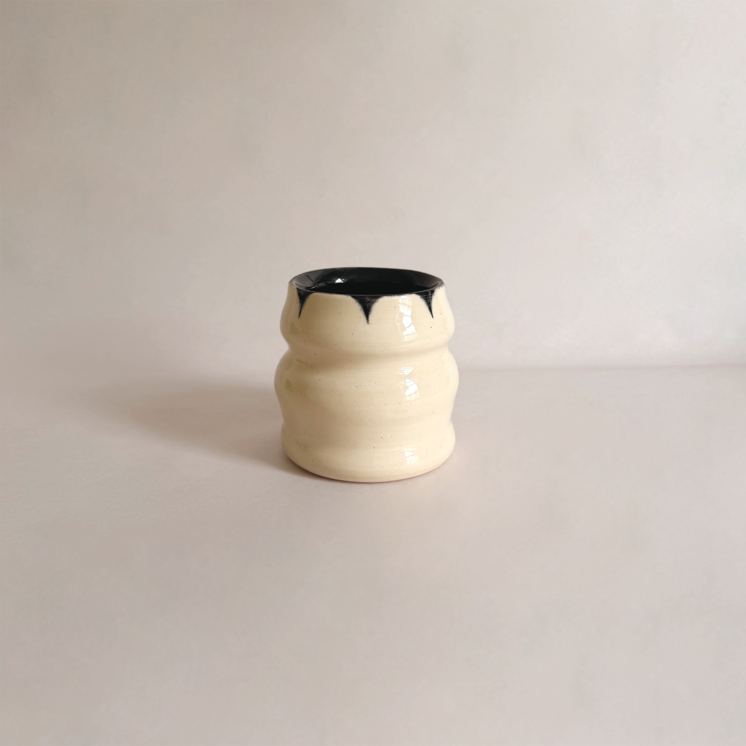 Cream-colored ceramic vase with black dripping glaze at the top, placed on a white surface with a plain white background.
