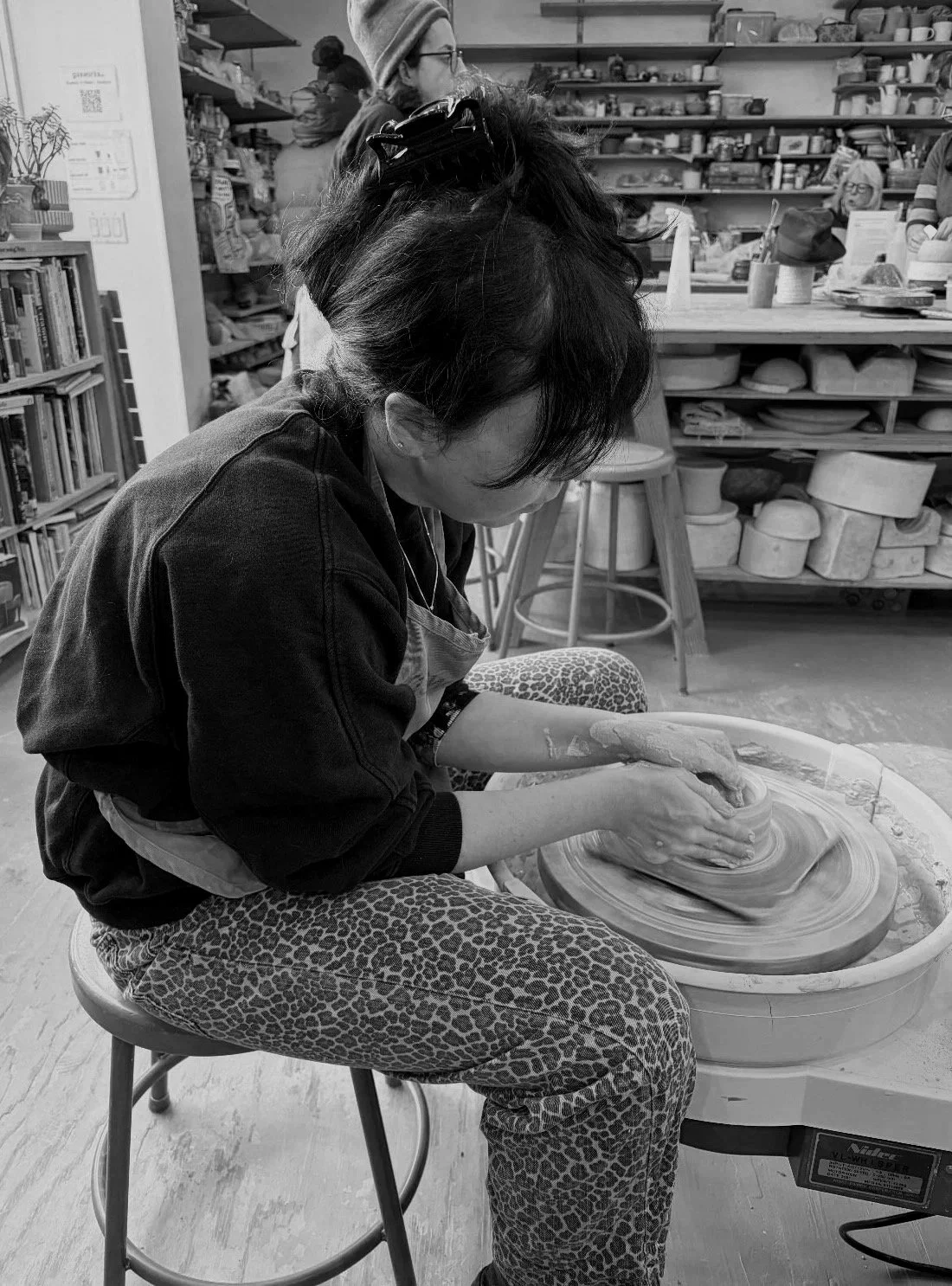 A woman is sitting on a stool and using a pottery wheel to shape clay in a ceramics studio. She is wearing leopard print pants and an apron, with her hair clipped back.