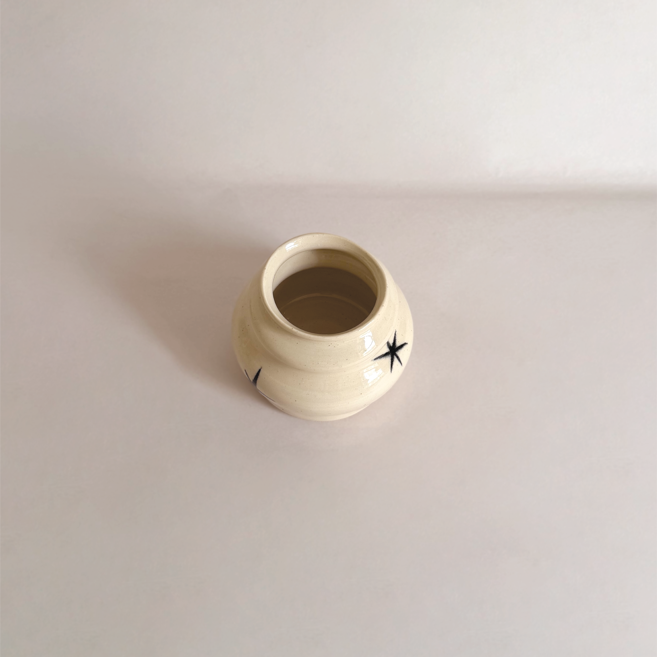Small cream-colored ceramic vase with black star and branch designs, viewed from above on a white surface.