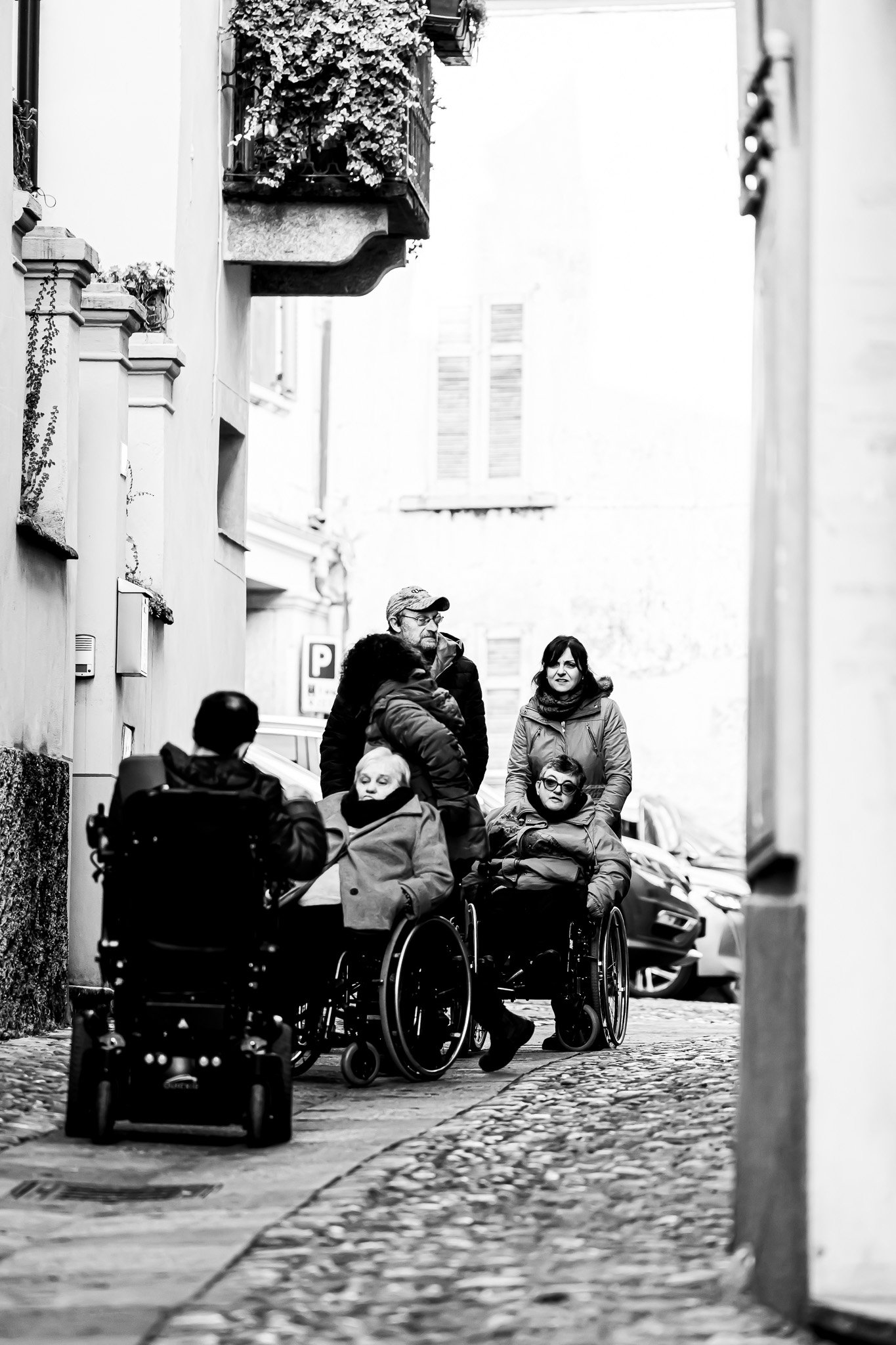 Persone anziane in carrozzina che si incontrano in una strada acciottolata di un quartiere residenziale, con edifici con balconi e finestre con persiane.