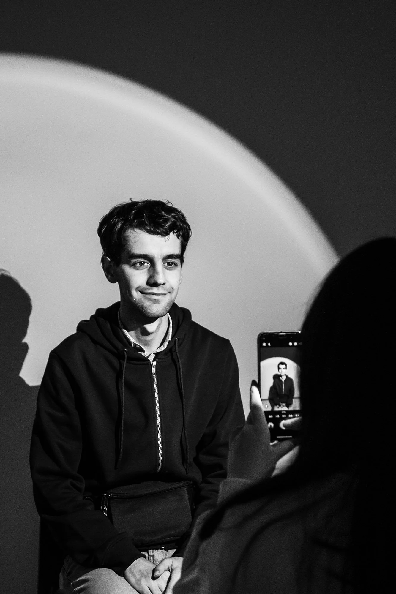 Un giovane uomo con capelli ricci, smilzo, indossa una felpa nera e sorride mentre una persona davanti a lui lo fotografa con uno smartphone. La scena è in bianco e nero.