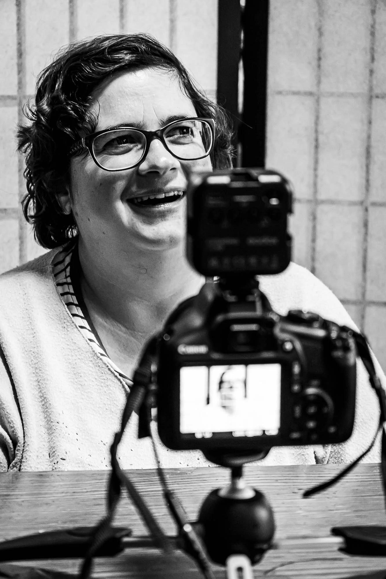 Ragazza con occhiali che sorride guardando verso la fotocamera, mentre si fotografa con una fotocamera digitale su un tavolo di legno, in bianco e nero.