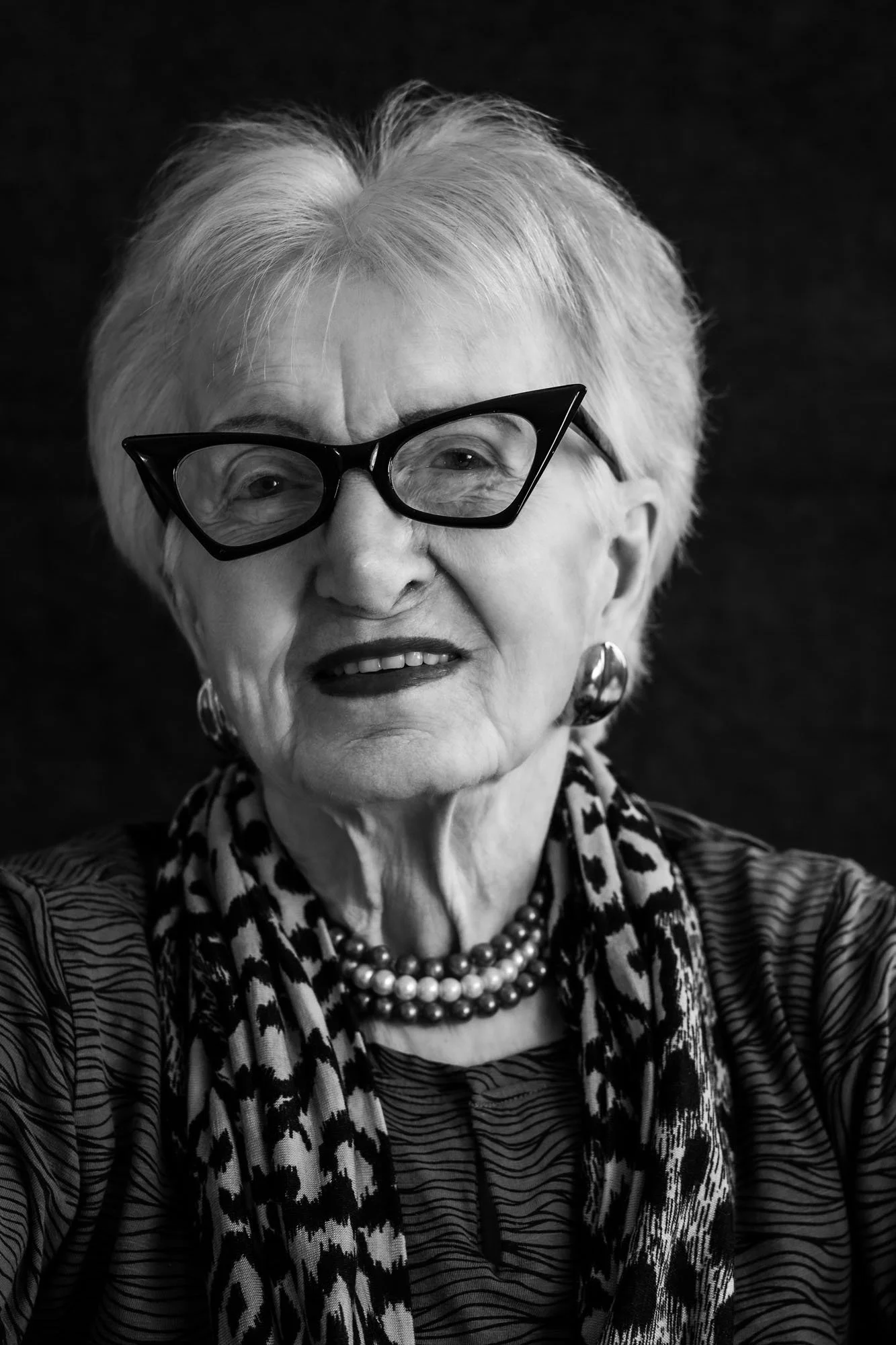 Foto in bianco e nero di una donna anziana che sorride, indossa occhiali di forma unica, orecchini grandi, collana di perle e una sciarpa con motivo leopardato, con sfondo nero.