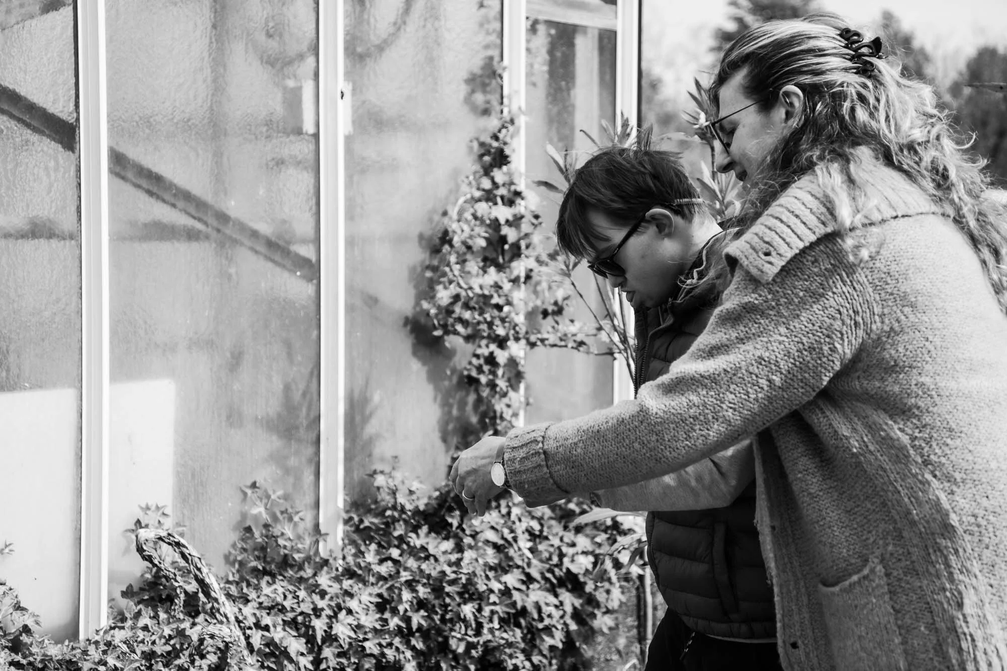 Due persone, una donna e un ragazzo, si chinano davanti a una finestra con vetri trasparenti, mentre osservano e fotografano le piante davanti a loro.