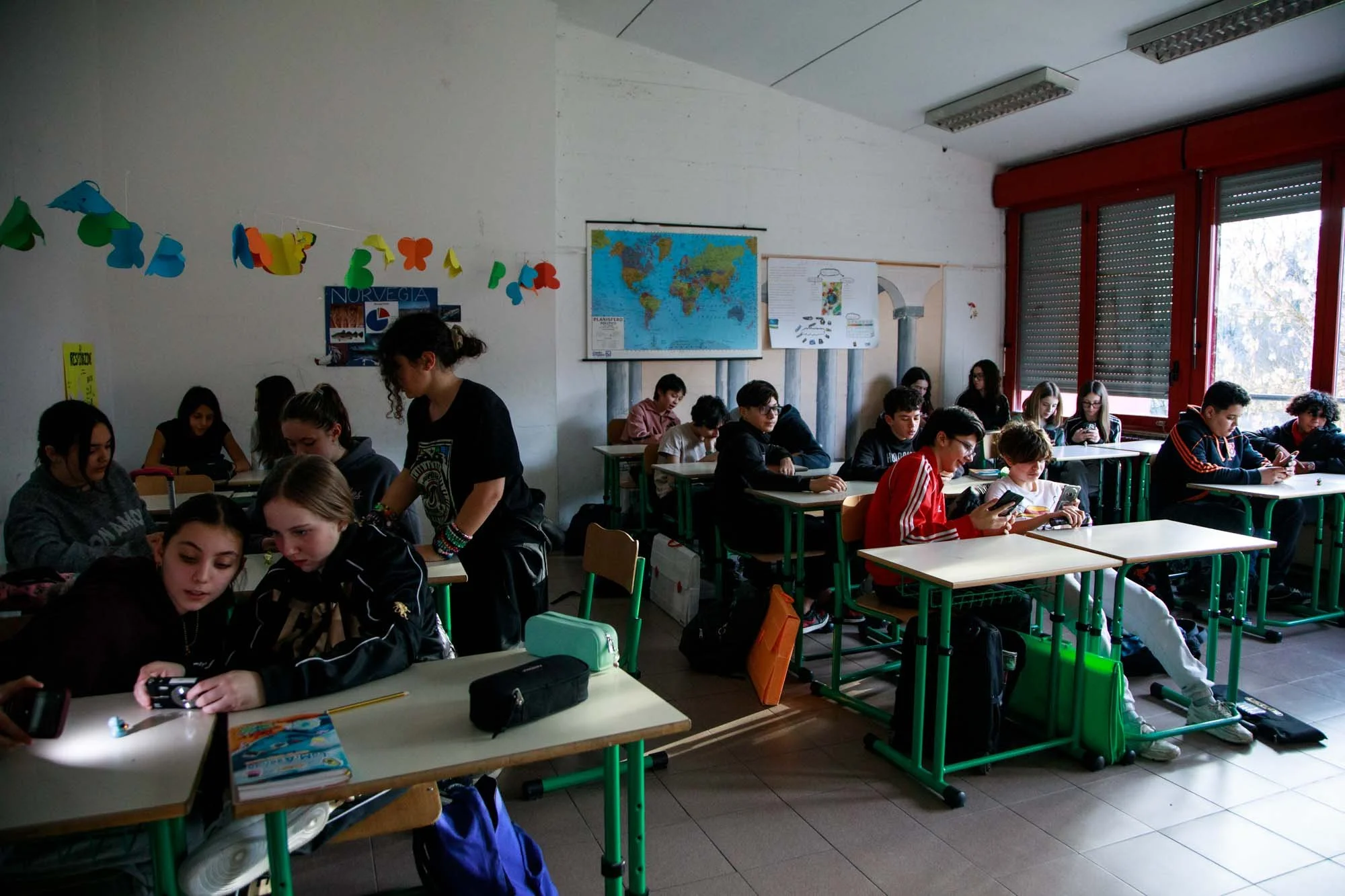 Classe di studenti in una scuola, alcuni usano il telefono cellulare, altri sono seduti a scrivere o ascoltare. In aula ci sono poster e mappe alle pareti, e grandi finestre.