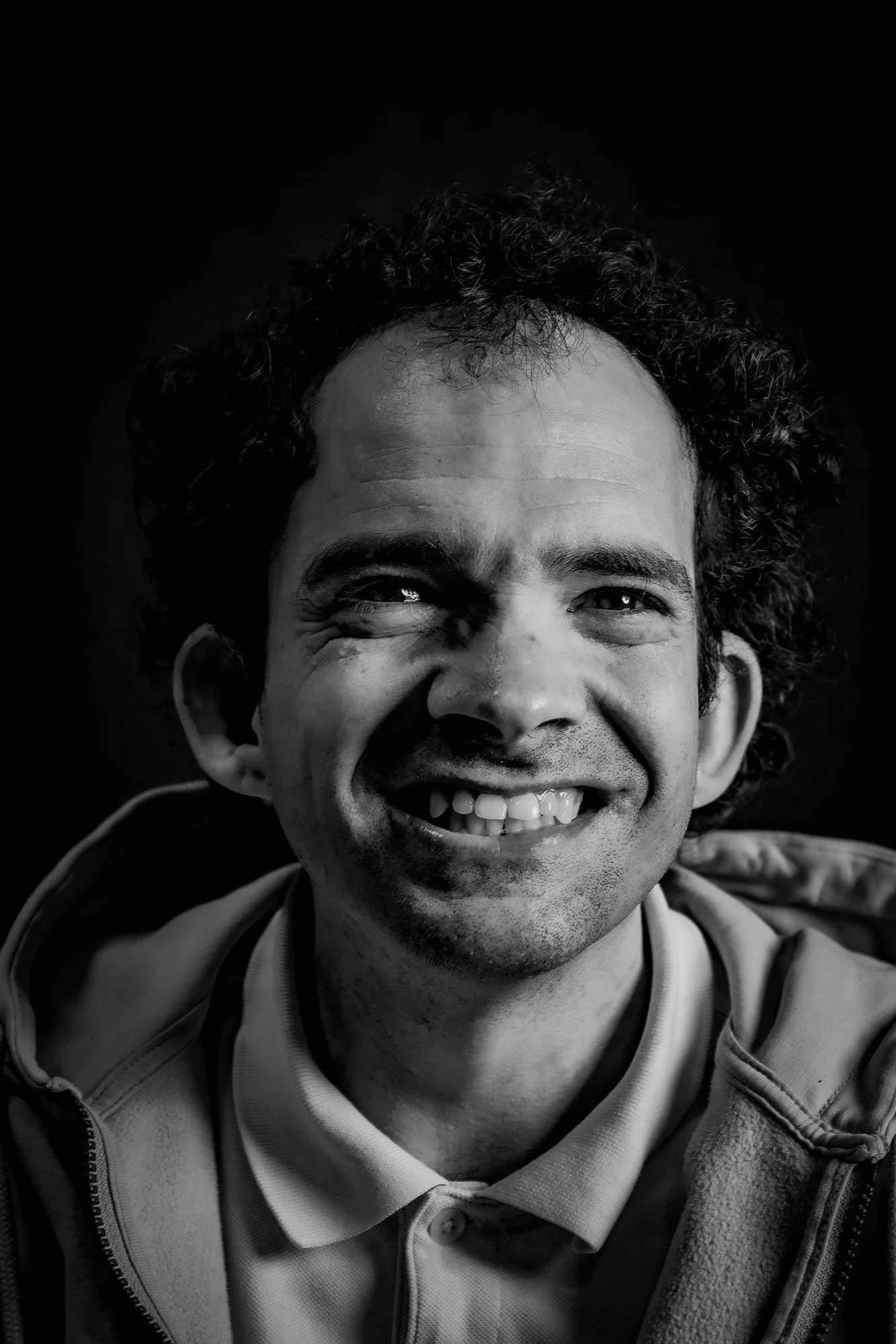 Ritratto in bianco e nero di un uomo con capelli riccioli e sorriso ampio, indossa una camicia e una giacca, sfondo nero.