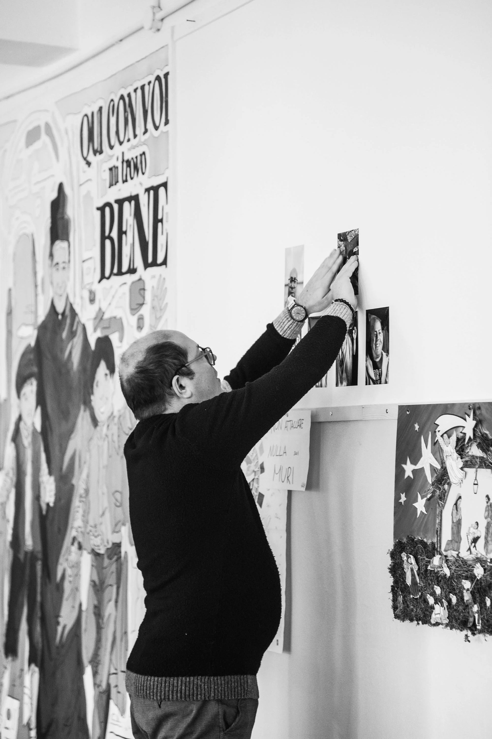 Uomo con occhiali appende fotografie al muro in una mostra o galleria, decorata con poster e disegni colorati.
