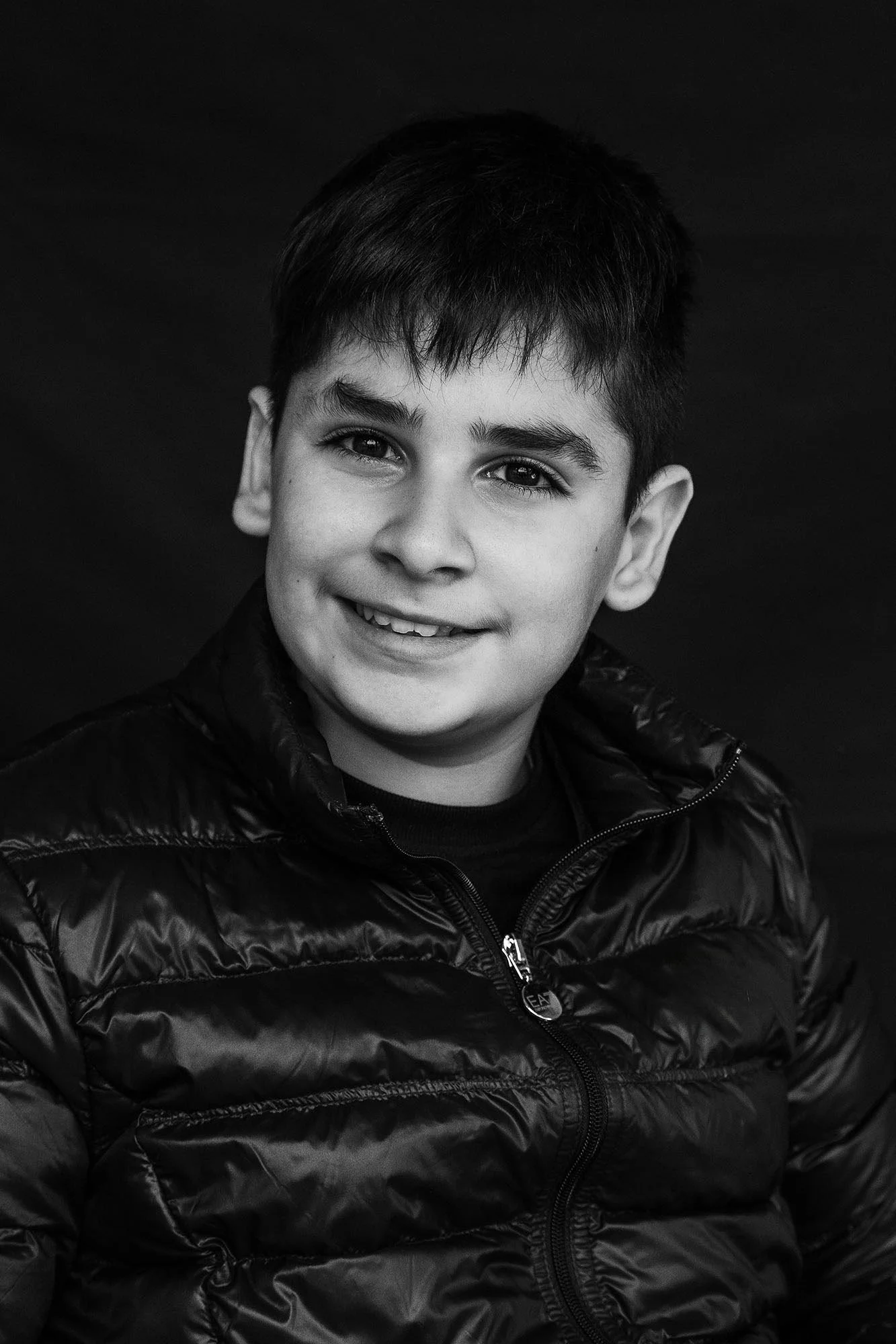 Ragazzo sorridente con capelli corti e scuri, indossa una giacca nera in nylon, sfondo nero.