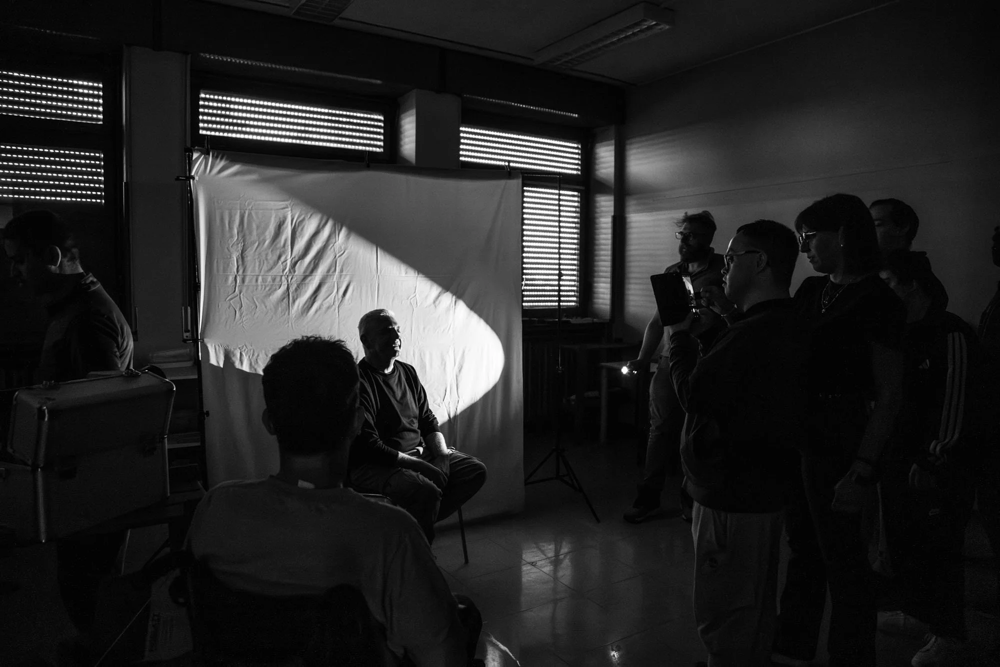 Scene di un gruppo di persone in una stanza buia, un uomo è seduto davanti ad un fondale bianco e illuminato dalla luce di una torcia, mentre i presenti lo fotografano con il loro cellulare.