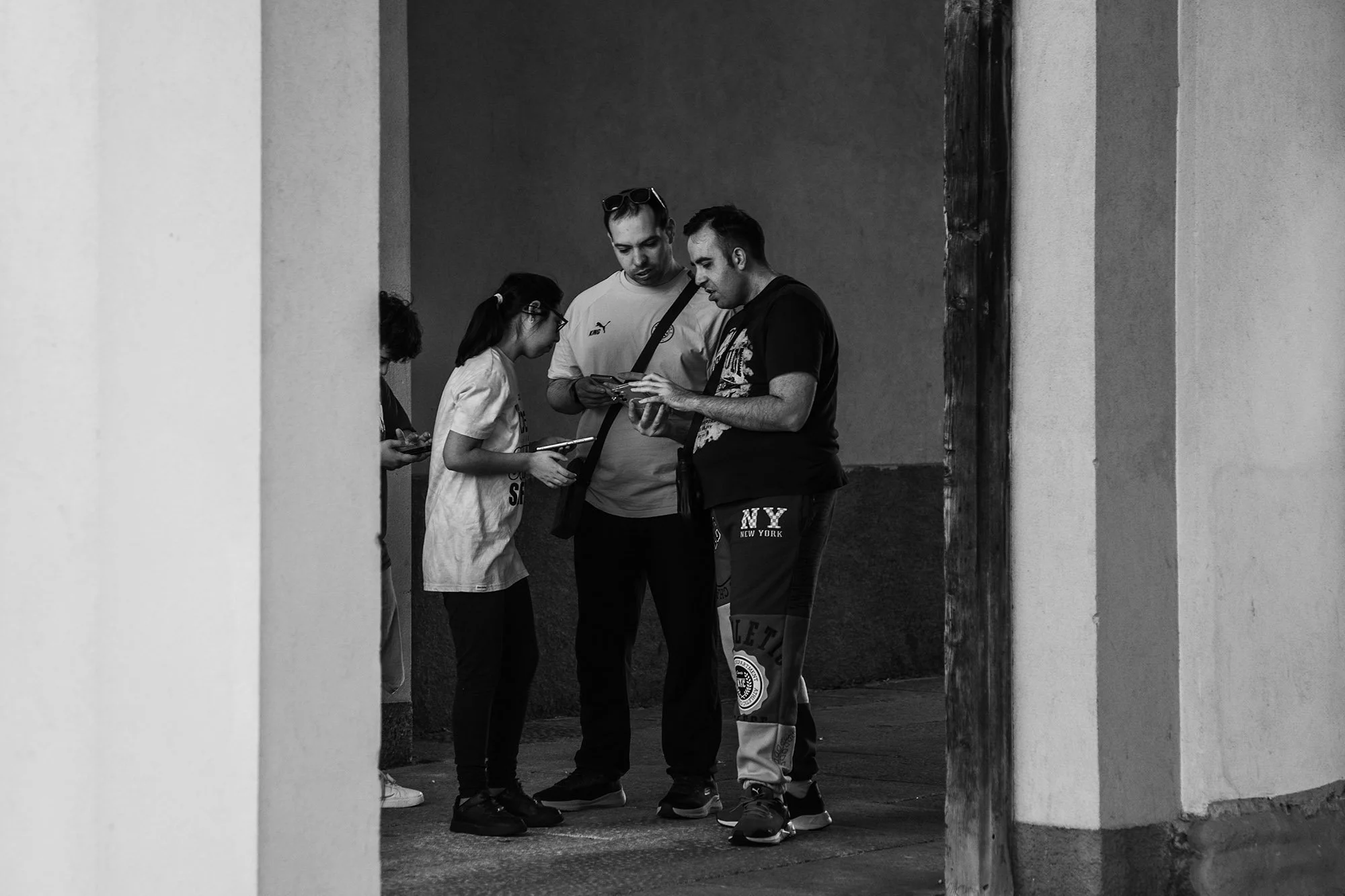 Tre persone si riuniscono in un corridoio, guardando uno smartphone, in un'immagine in bianco e nero.