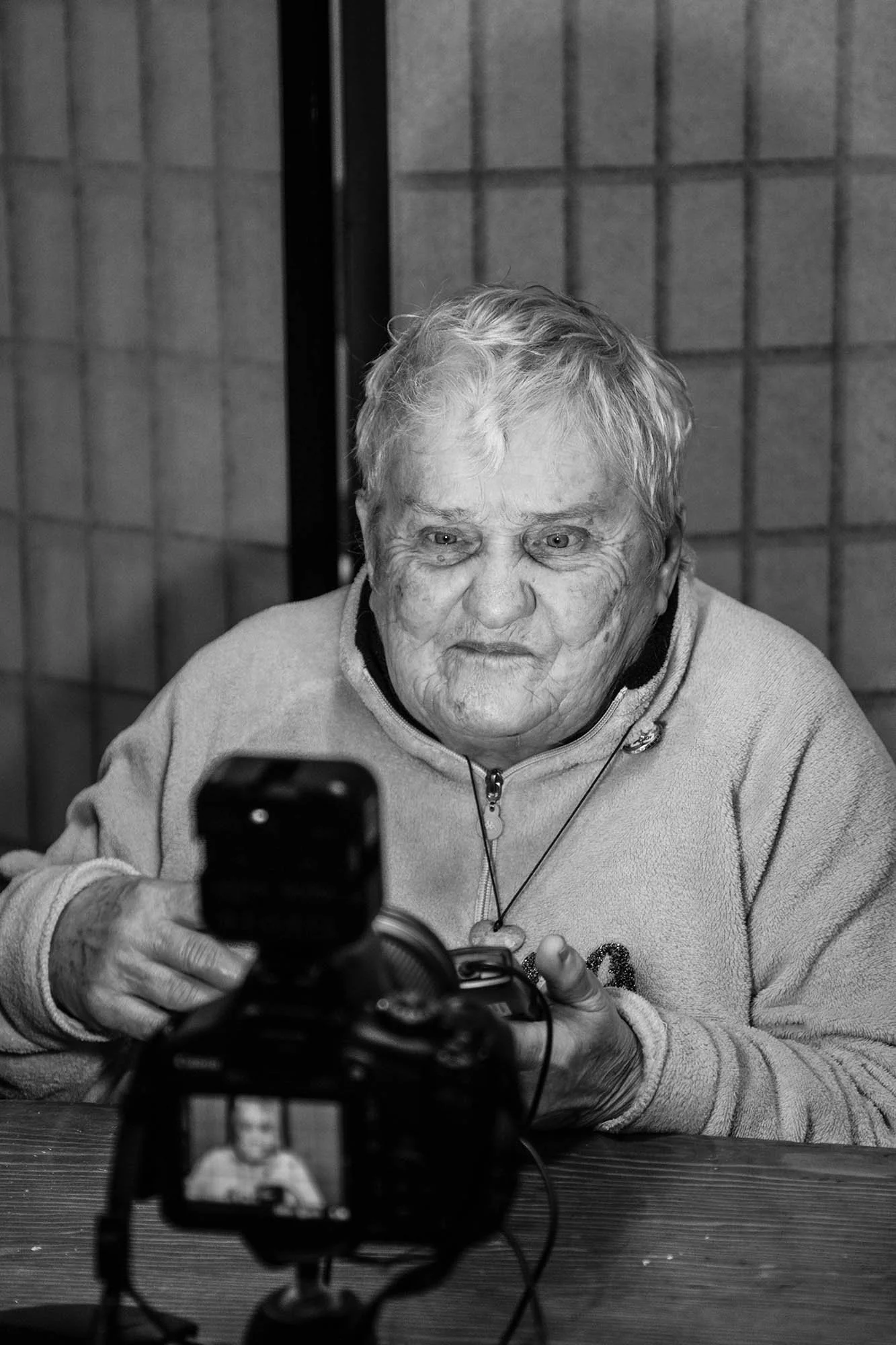 Una donna anziana seduta a una tavola, con una fotocamera davanti a lei che registra o scatta una foto.