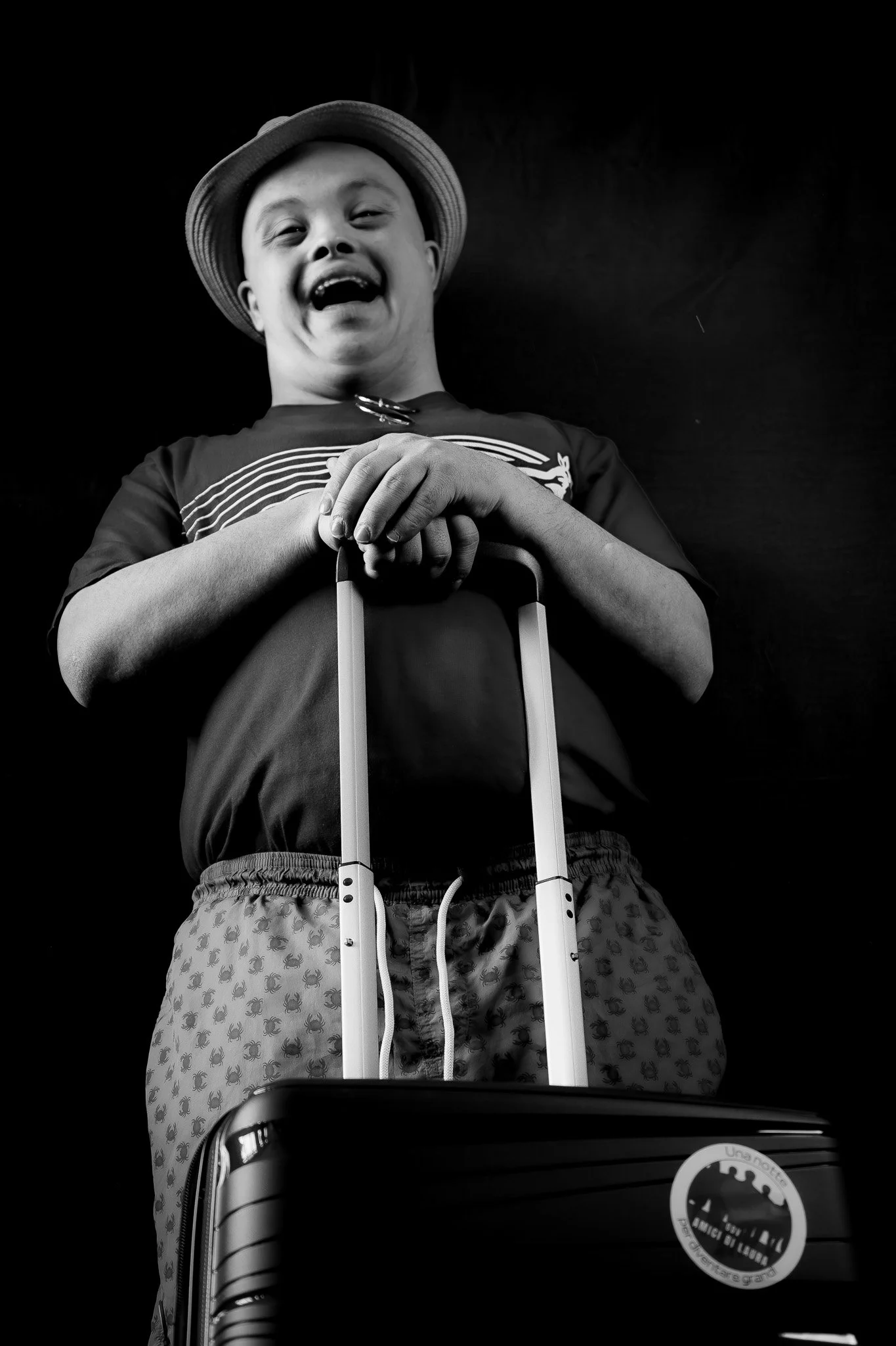 Bambino sorridente con cappello, maglietta a righe e pantaloni con motivo di pappagalli, che tiene una maniglia di valigia e un bagaglio a mano in primo piano, scena in bianco e nero.