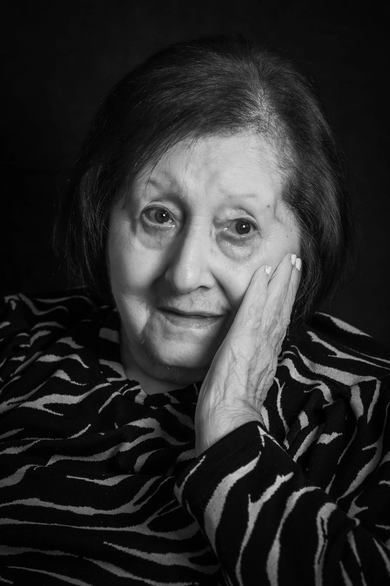 Ritratto di una donna anziana con capelli scuri e occhi sorridenti, che si copre parte del volto con una mano, indossa una maglietta zebrata. Fotografia in bianco e nero.