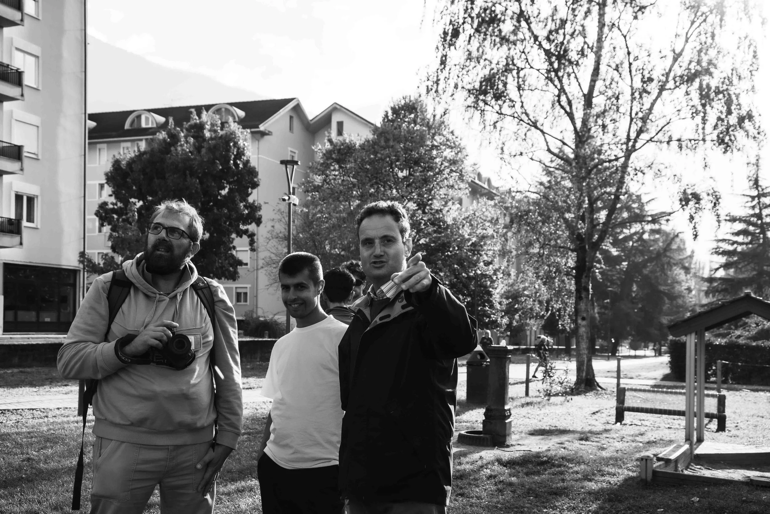 Un gruppo di tre uomini in un parco cittadino, alcuni sorridenti e uno che indica qualcosa verso la fotocamera, con alberi e edifici sullo sfondo, in bianco e nero.