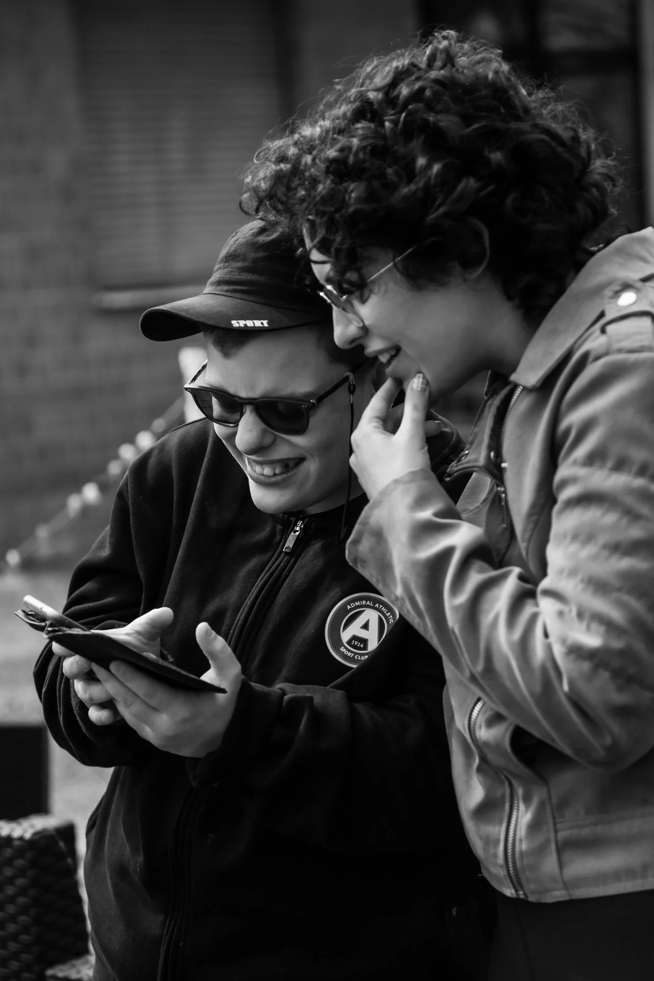 Due persone sorridenti, una con cappellino e occhiali da sole, l'altra con capelli ricci e occhiali, guardano uno smartphone insieme.