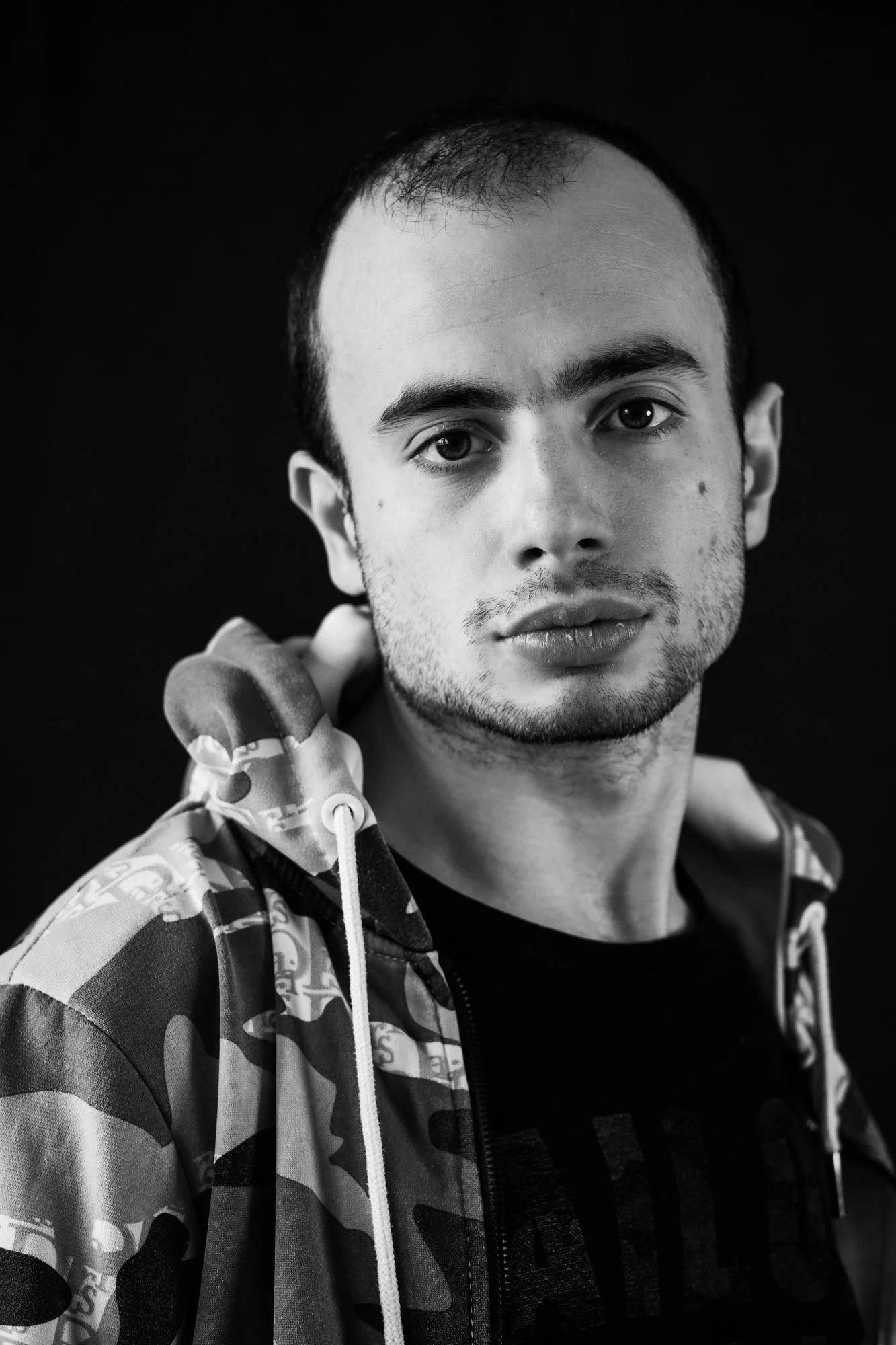 Ritratto di un giovane uomo che tiene un manubrio da mano a spalla, veste una felpa con cappuccio con disegni militari, sfondo nero.