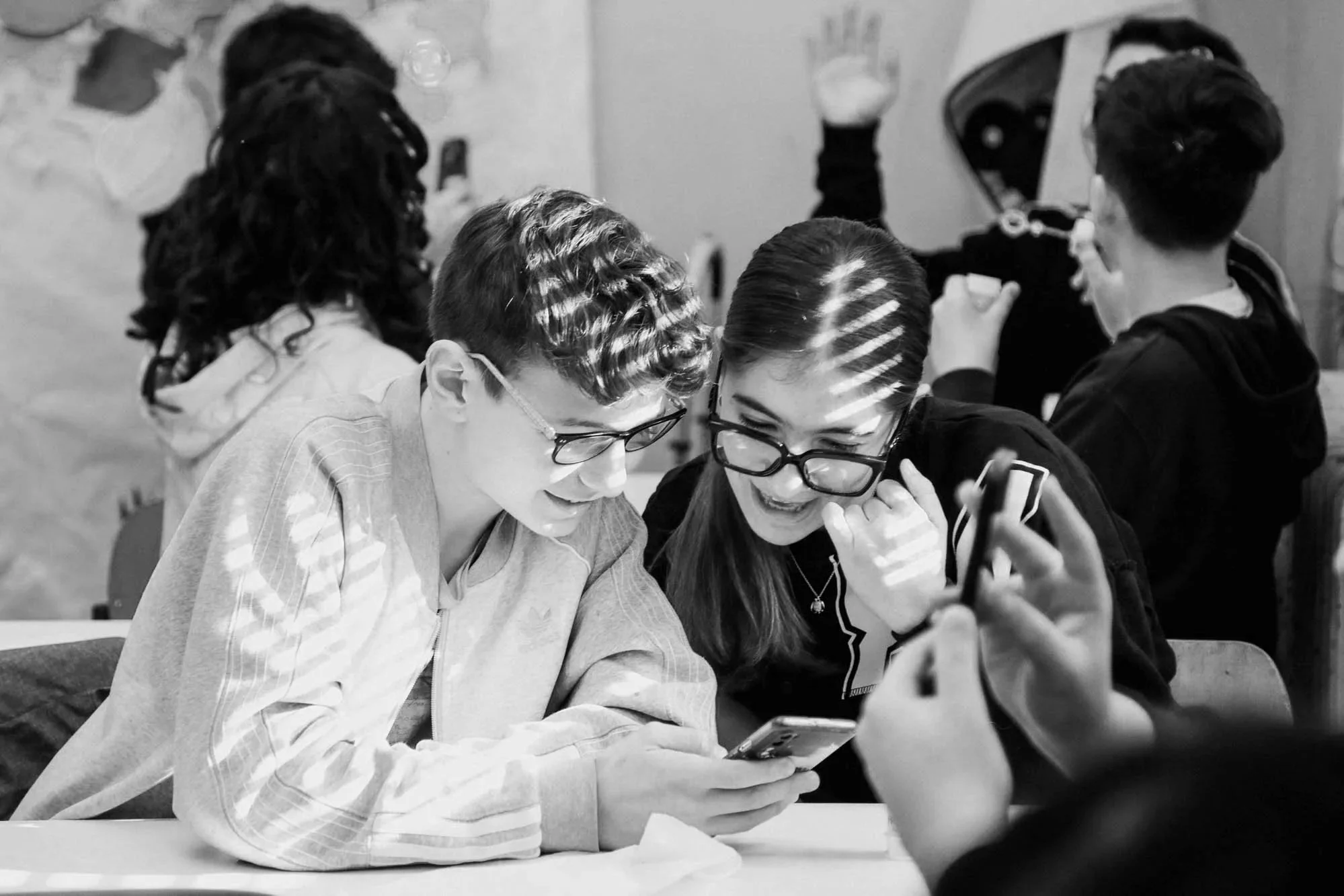Due giovani donne si scambiano un sorriso mentre guardano uno smartphone, sedute a un tavolo in un ambiente di gruppo.