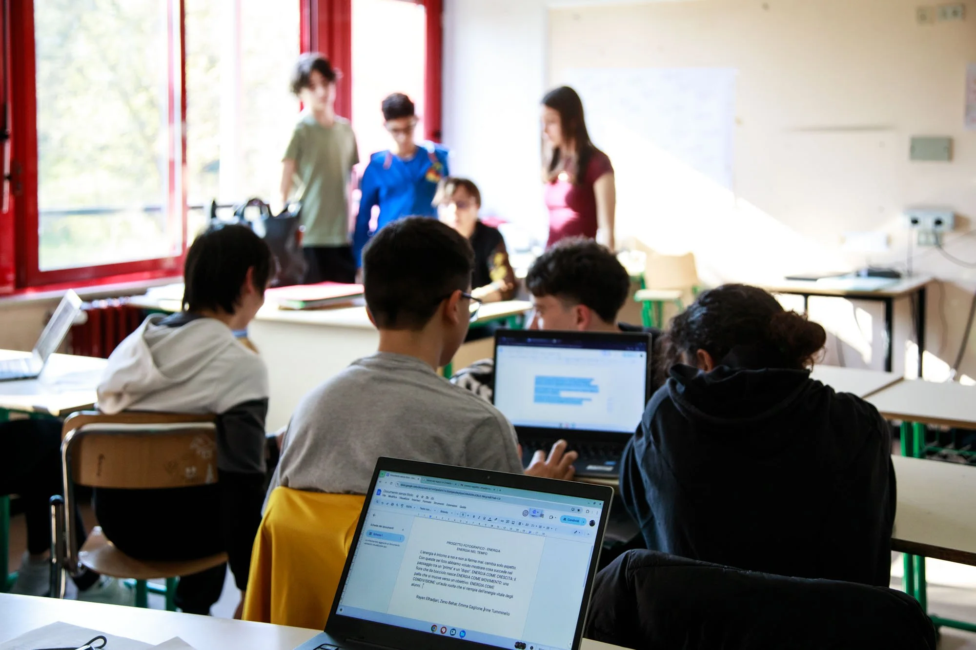 Classe scolastica con studenti seduti ai rispettivi tavoli e laptop, insegnante che parla, finestra con luce naturale.