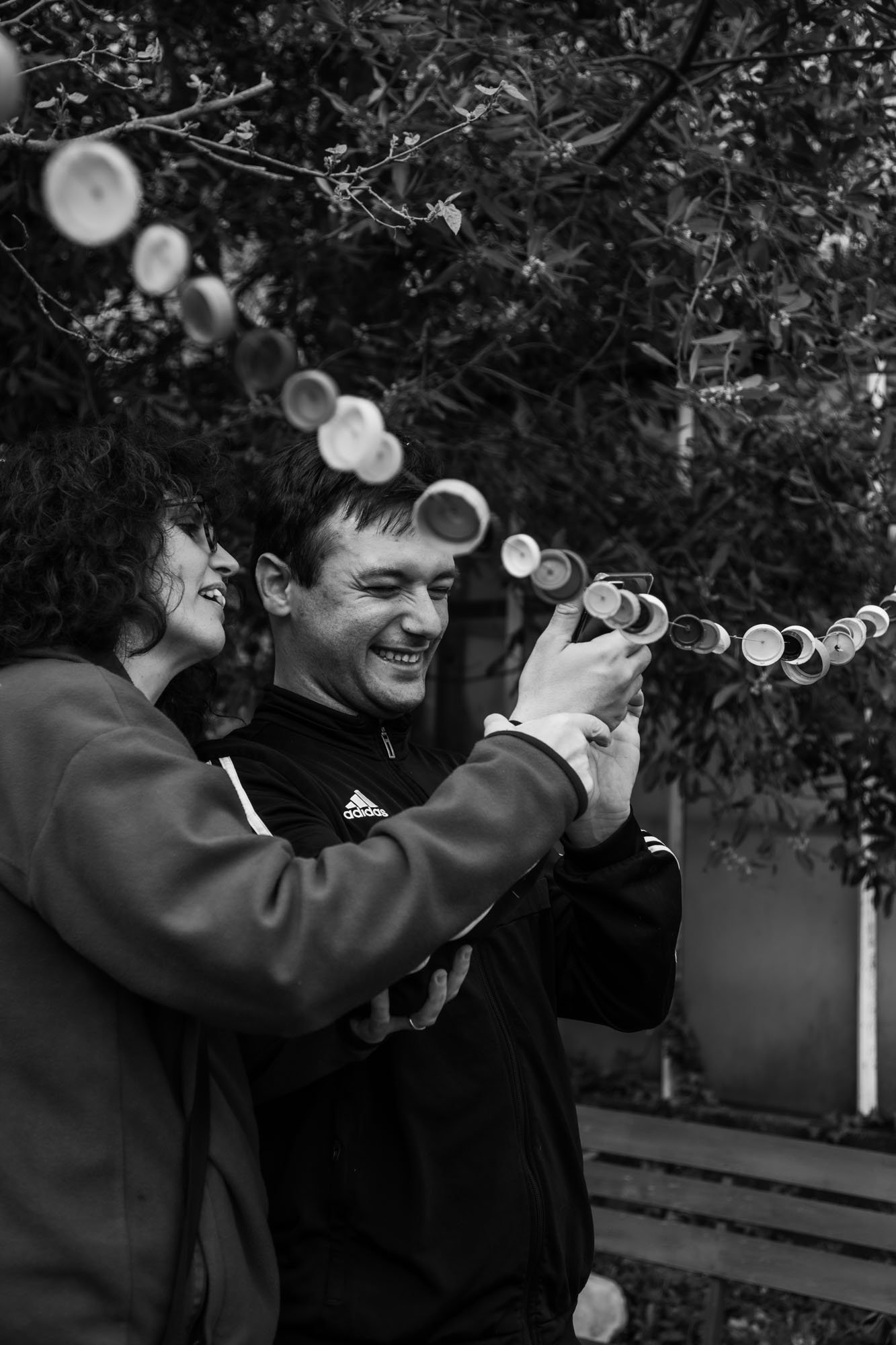 Due persone sorridenti, una donna e un uomo, scattano una foto con uno smartphone in un parco, circondati da arbusti e alberi.
