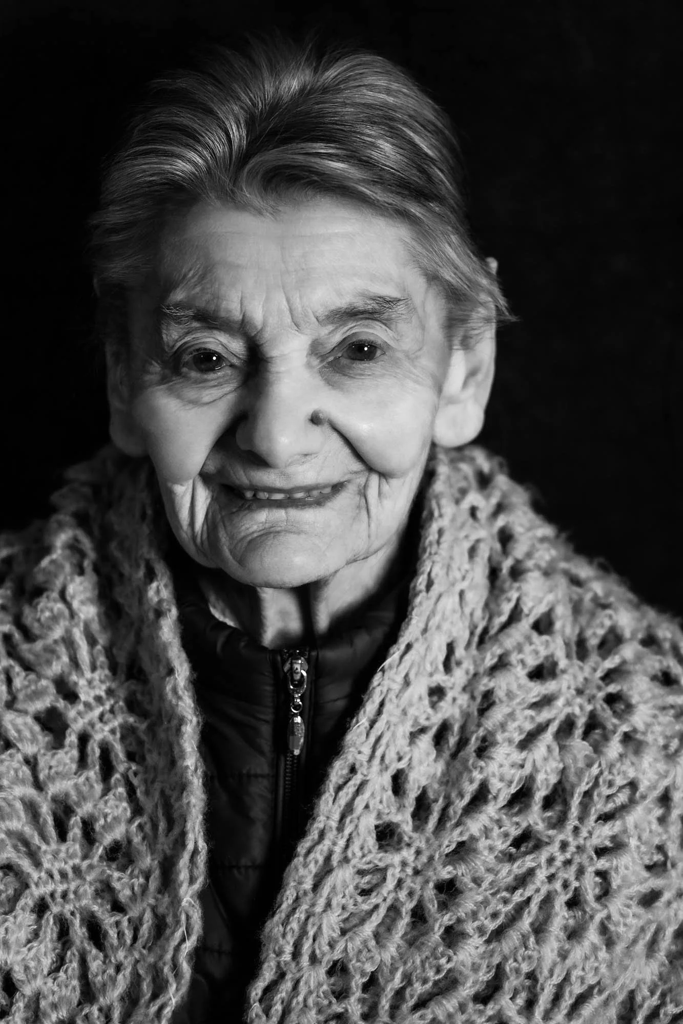 Foto in bianco e nero di una donna anziana con capelli raccolti, avvolta in uno scialle a maglia grossa, che sorride leggermente.