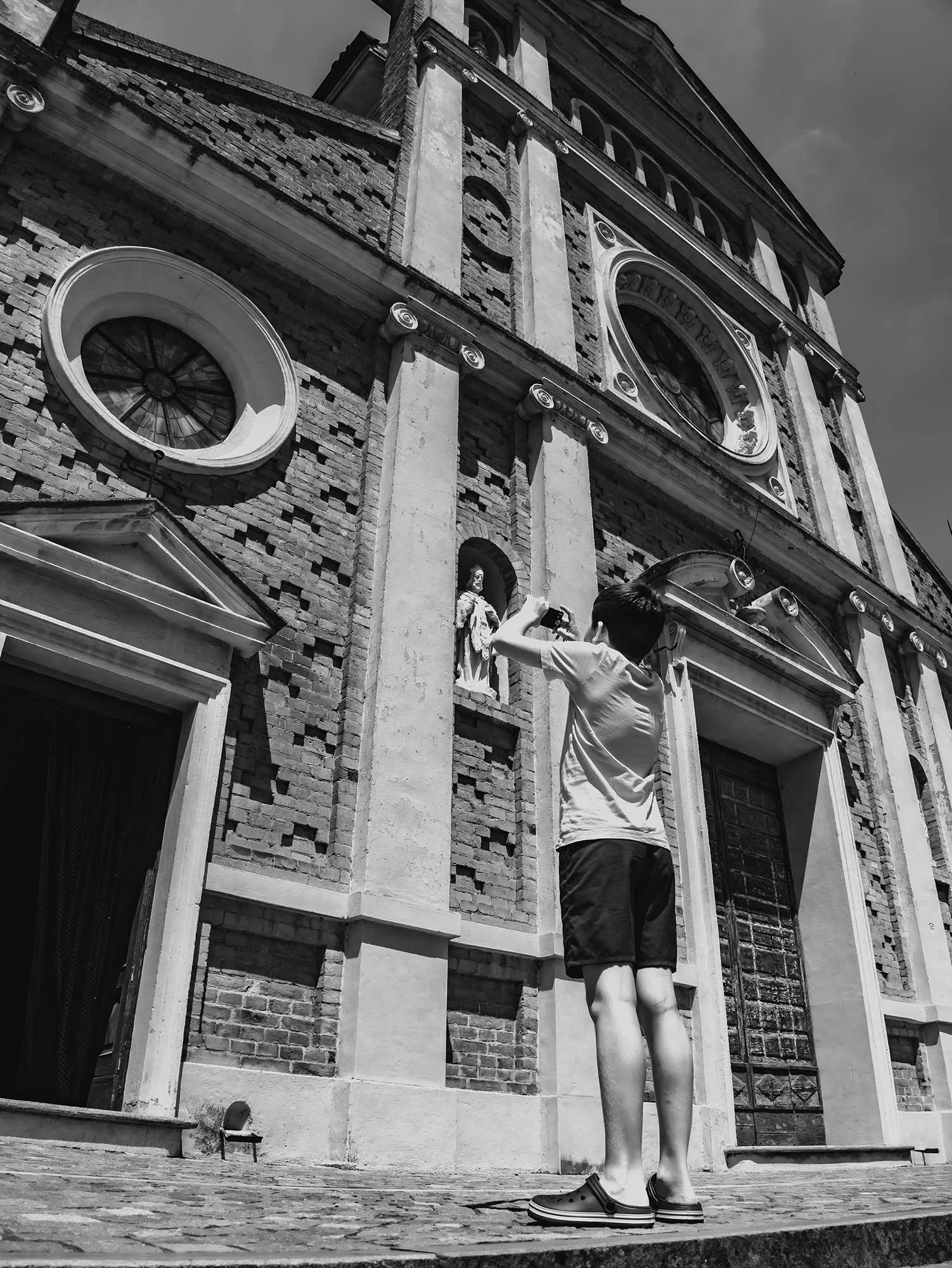 Un ragazzo con pantaloncini corti scatta una foto davanti ad una chiesa, con un grande rosone sulla facciata, in un giorno soleggiato.