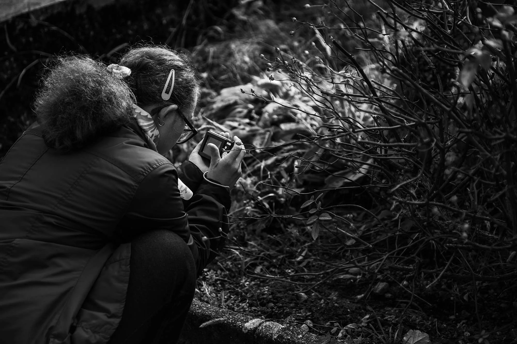 Una donna con capelli ricci e occhiali da vista prende una foto con una macchina fotografica digitale tra cespugli e natura.