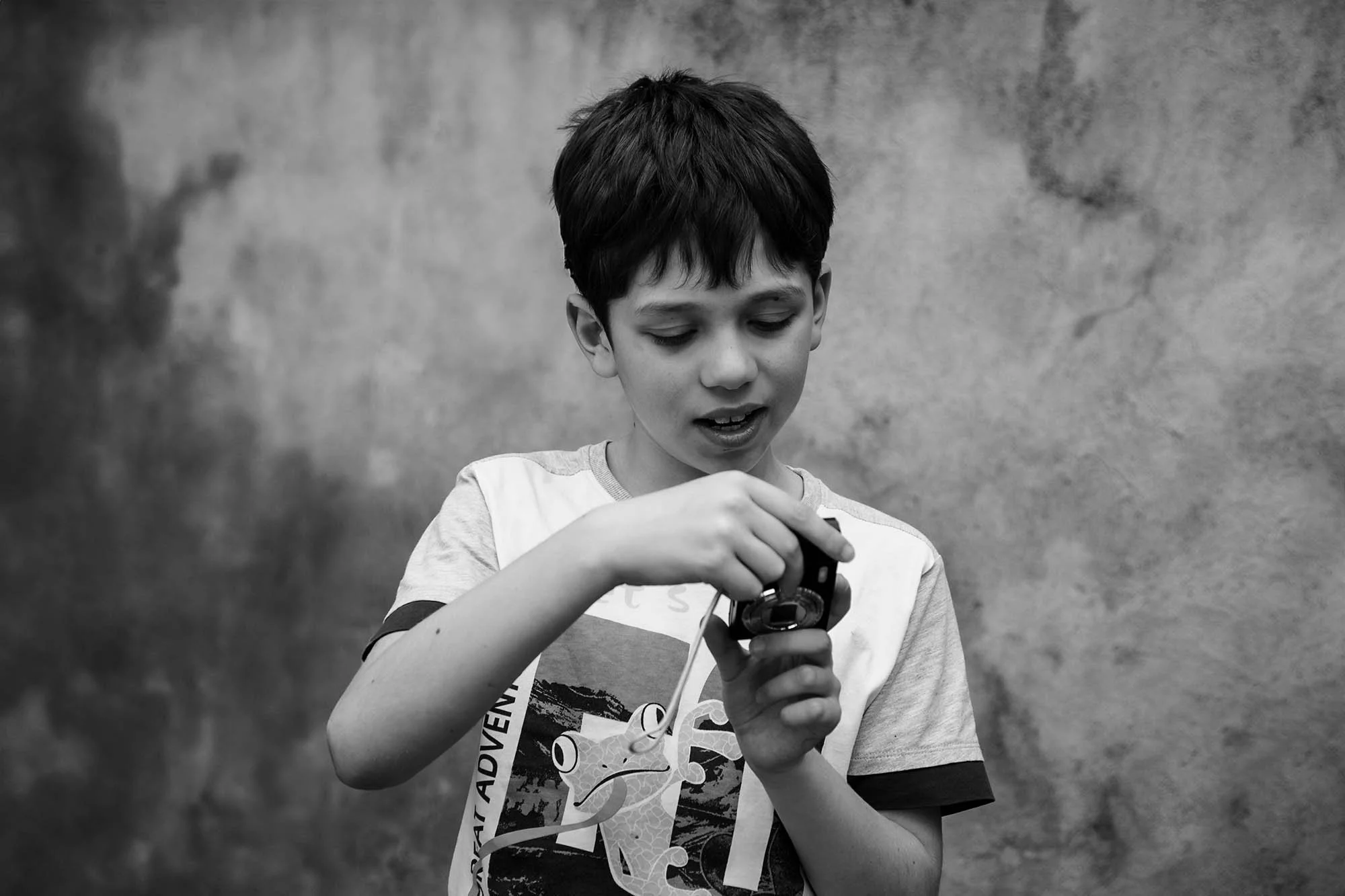 Ragazzo con capelli corti, vestito con maglietta bianca, che guarda lo schermo di una macchina fotocamera compatta, con uno sfondo di muro grigio.