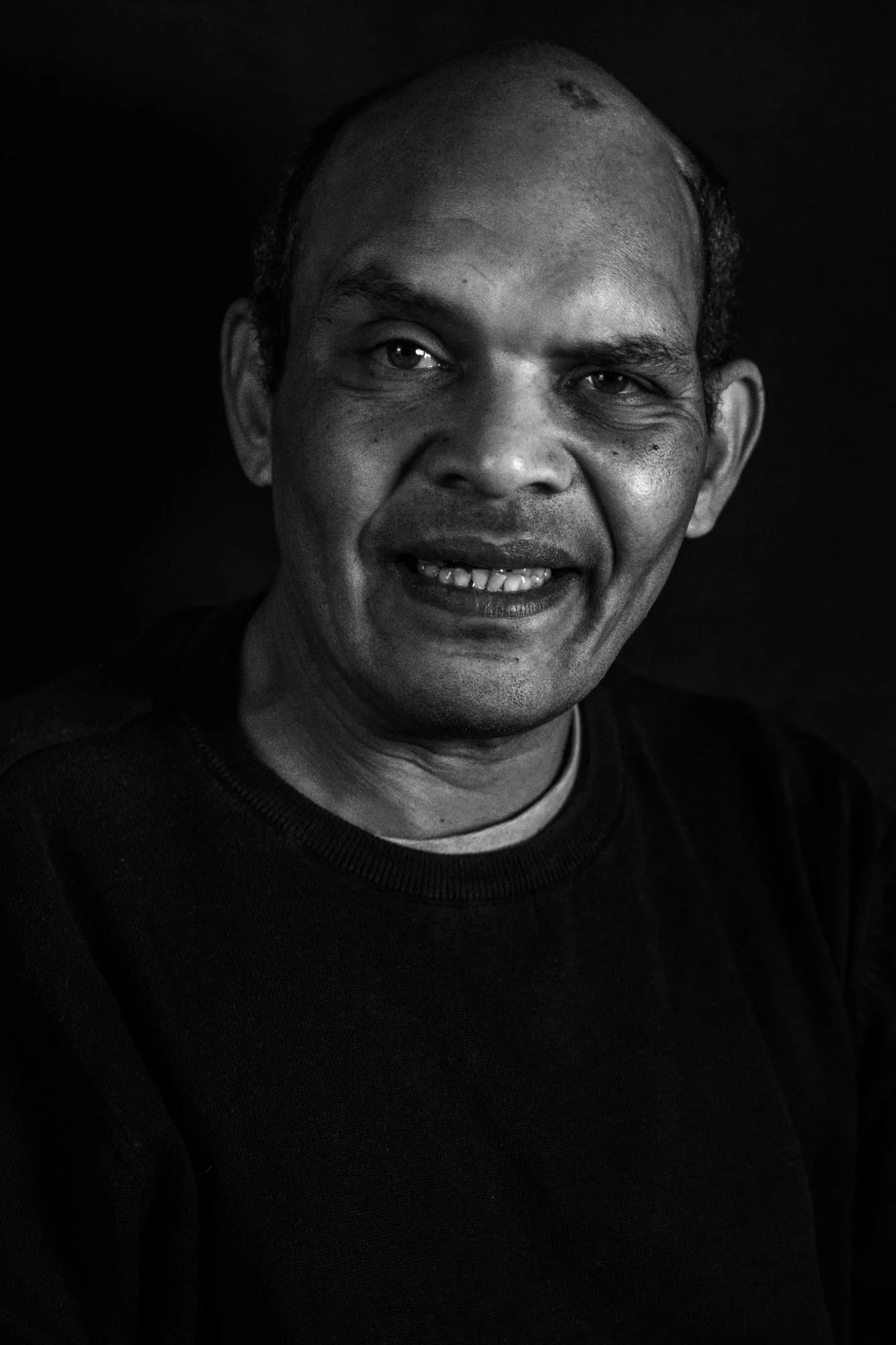 Foto in bianco e nero di un uomo di mezza età con pelle scura, capelli corti e sorridente, indossa una maglia nera.