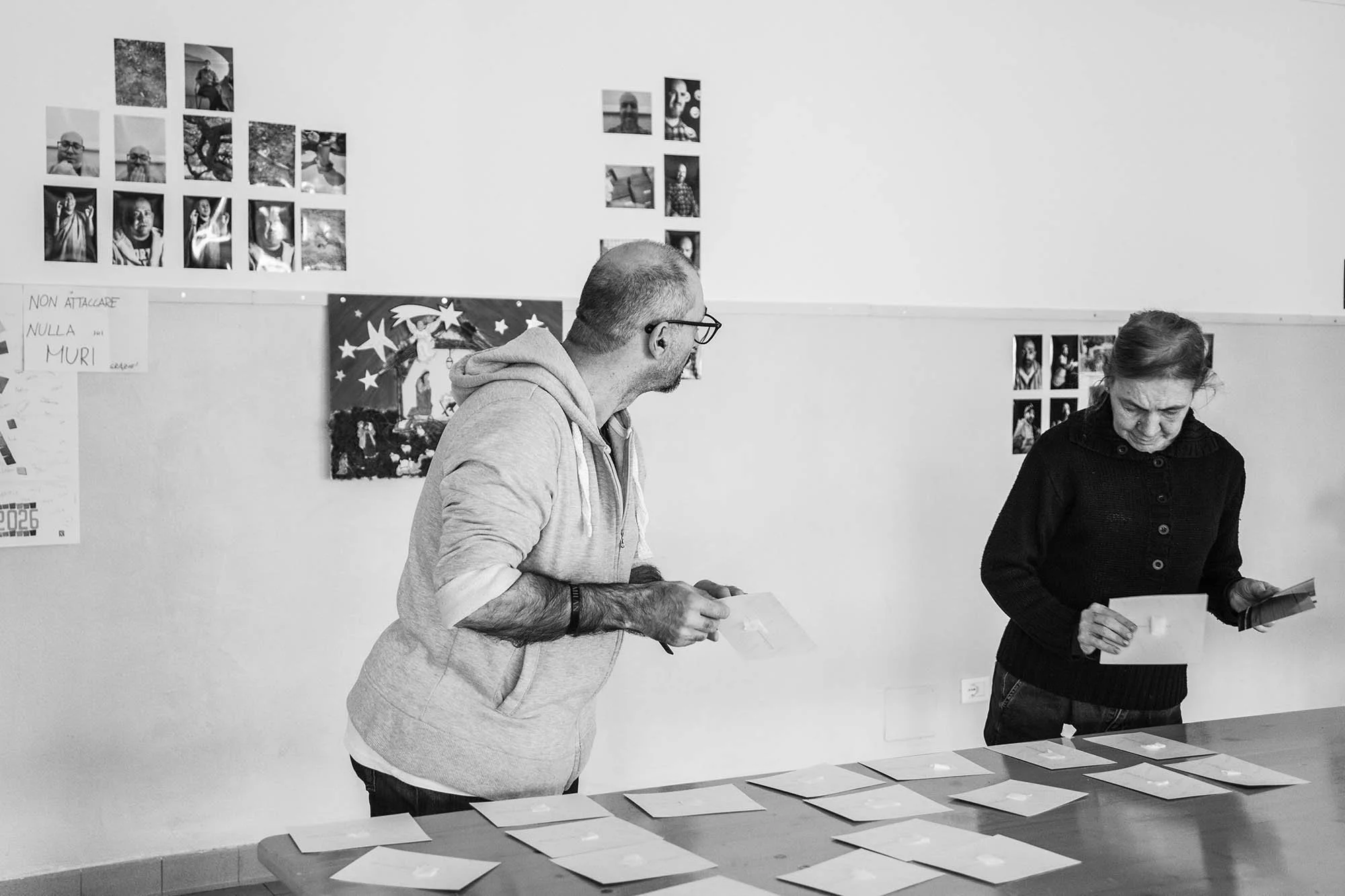 Due persone, un uomo e una donna, stanno esaminando delle fotografie su un tavolo. Sul muro dietro di loro ci sono diverse fotografie di persone e un poster con un angelo e stelle. L'ambiente sembra essere una sala espositiva o un laboratorio di arte