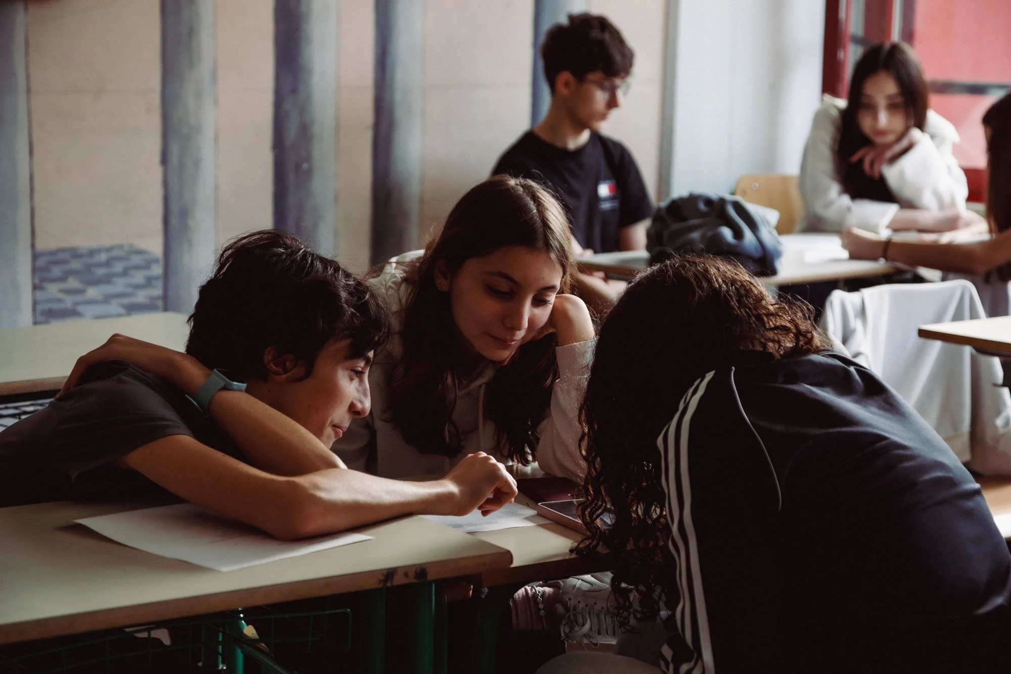 Gruppo di giovani studenti seduti a un banco di scuola, alcuni si confrontano mentre gli altri guardano e interagiscono.