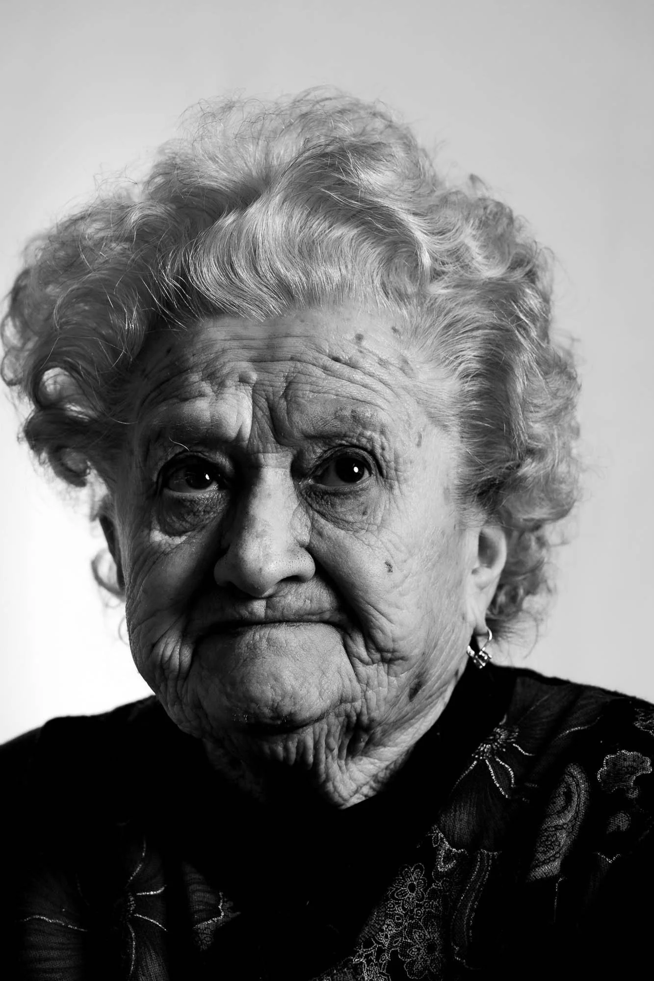 Ritratto in bianco e nero di una donna anziana con capelli ricci e faccia segnate da rughe, indossa orecchini e una maglia scura.