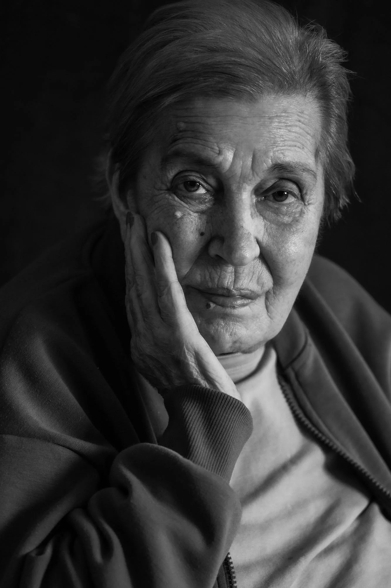 Ritratto in bianco e nero di una donna anziana con capelli grigi, che tocca il volto con una mano, esprimendo malinconia o riflessione.