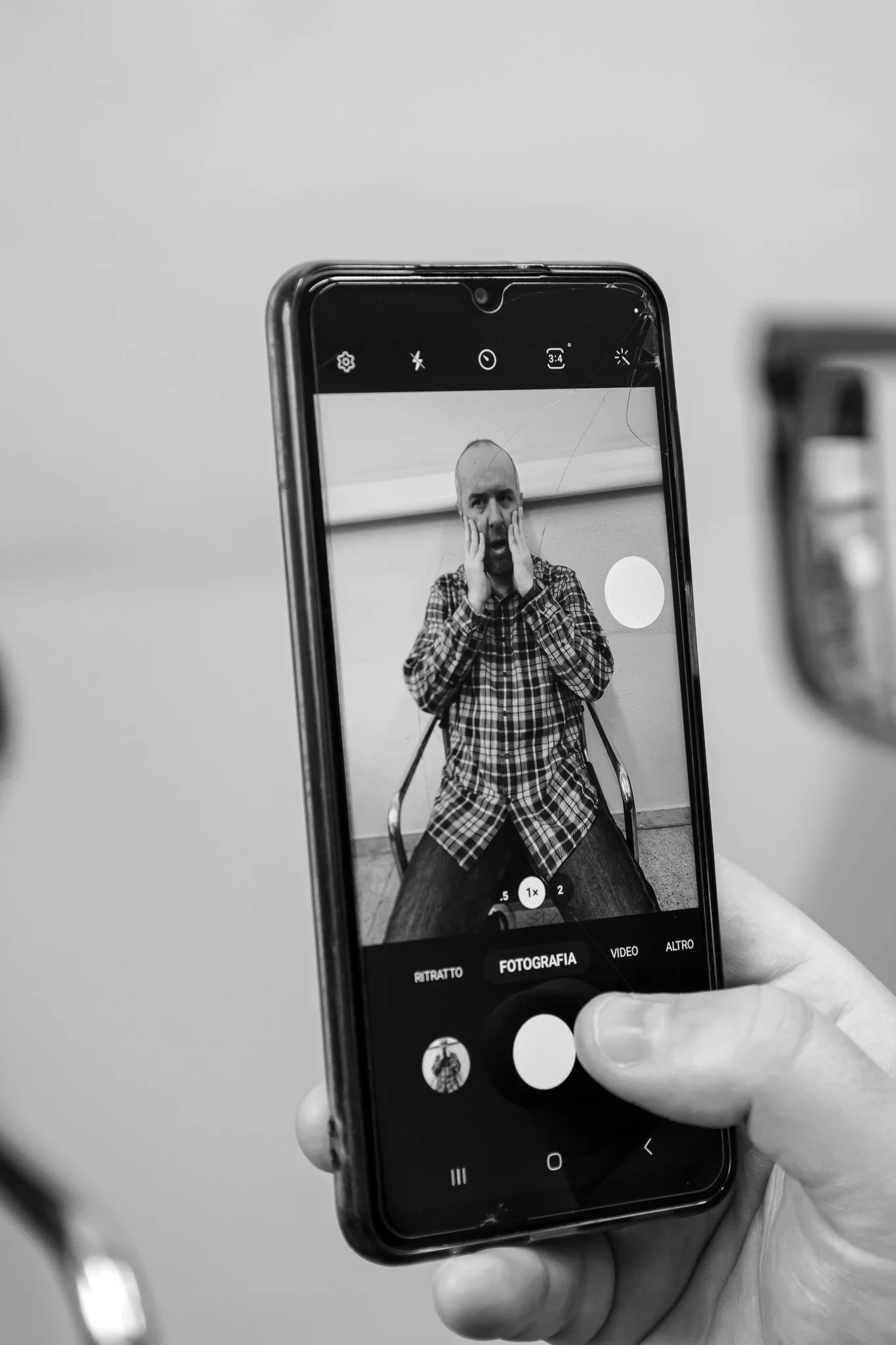 Una persona tiene il telefono cellulare e fotografa un uomo con le mani al viso. Il telefono mostra un'immagine in bianco e nero di lui, indossa una camicia a quadri e si trova in un ambiente chiuso.