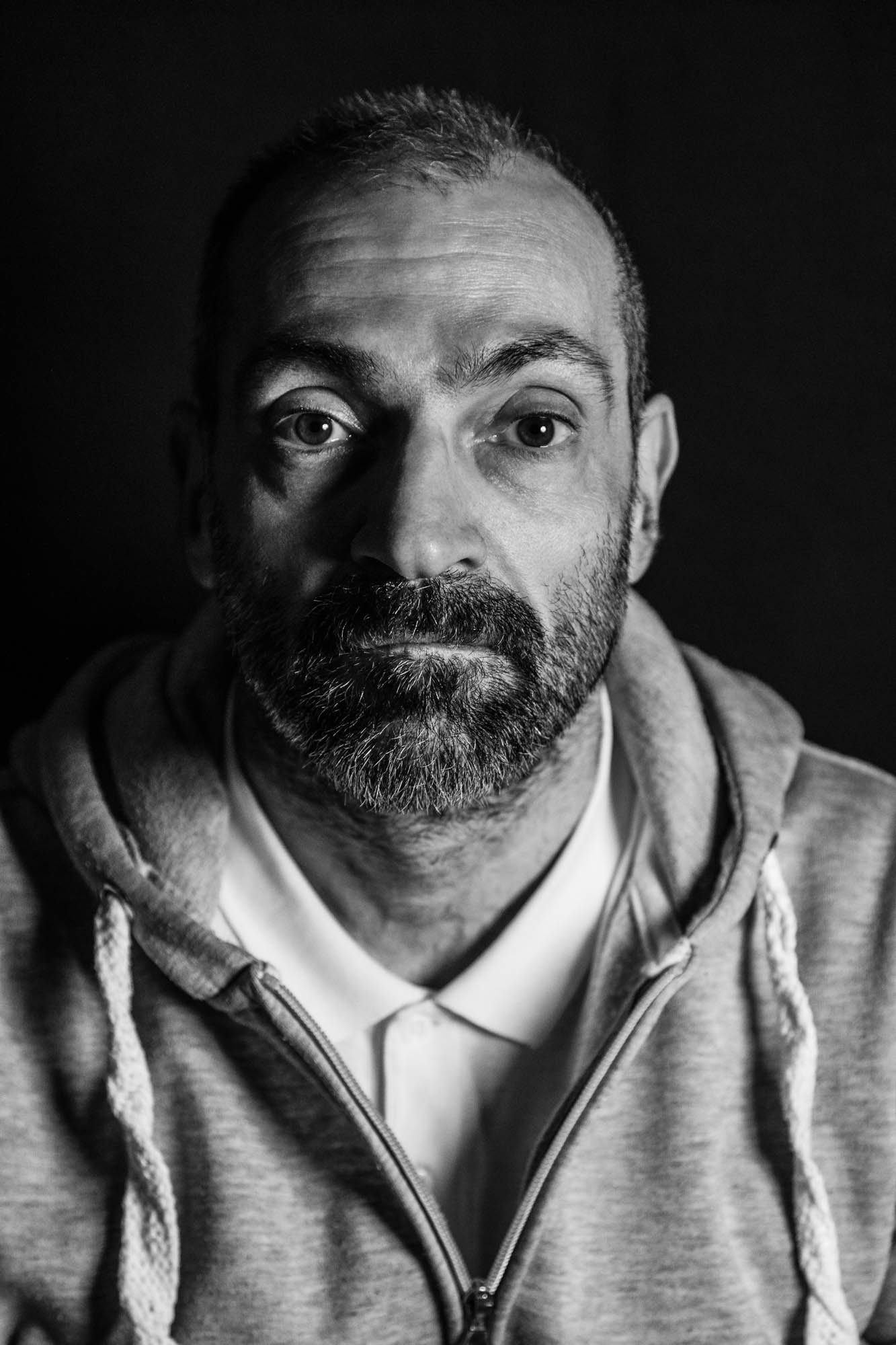 Uomo con barba e capelli corti, vestito con felpa grigia con cappuccio e zip, sfondo nero.