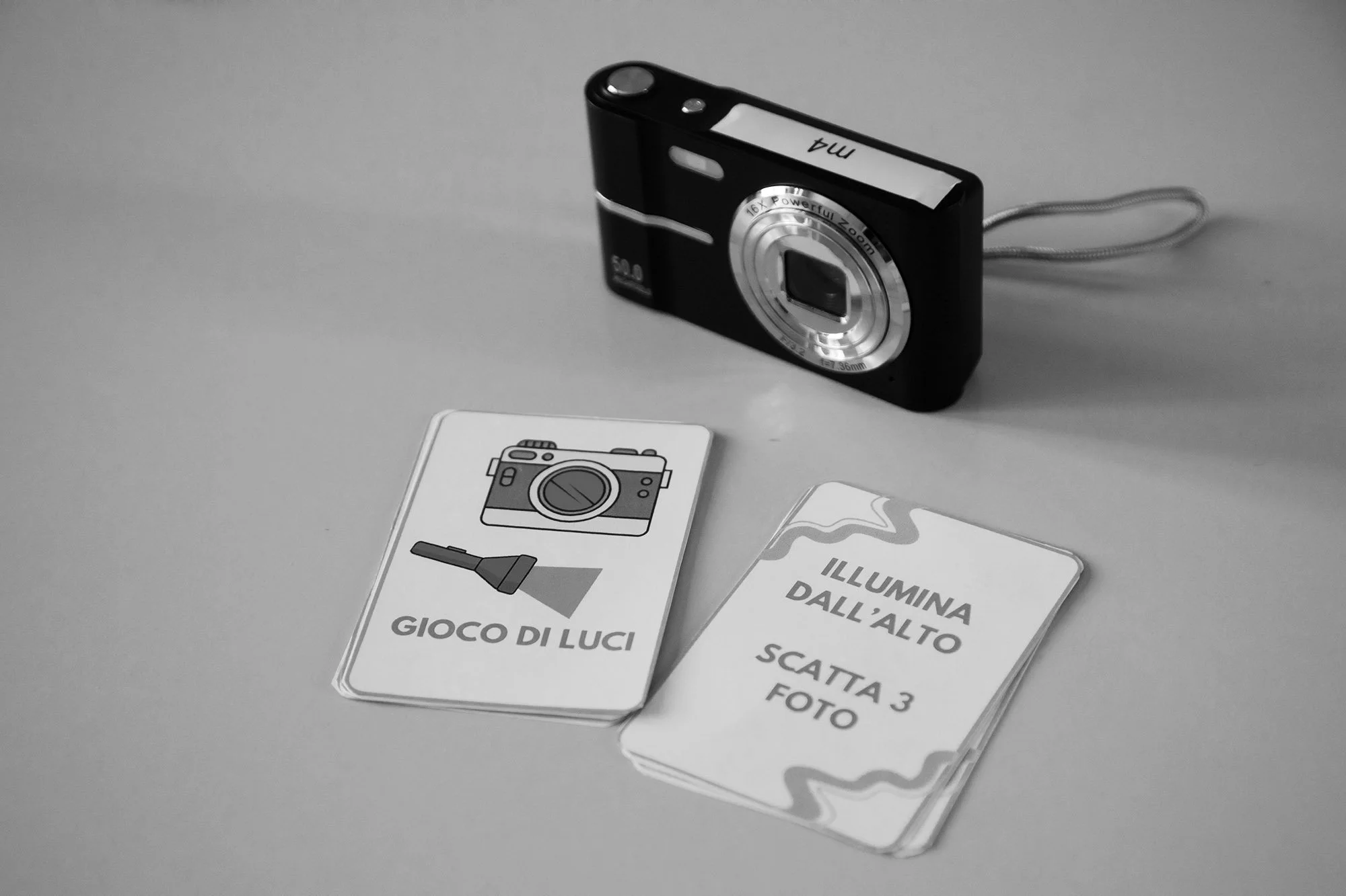 Fotocamera digitale nera con precamera argentata e cinturino, carte con istruzioni in italiano e immagine di una fotocamera