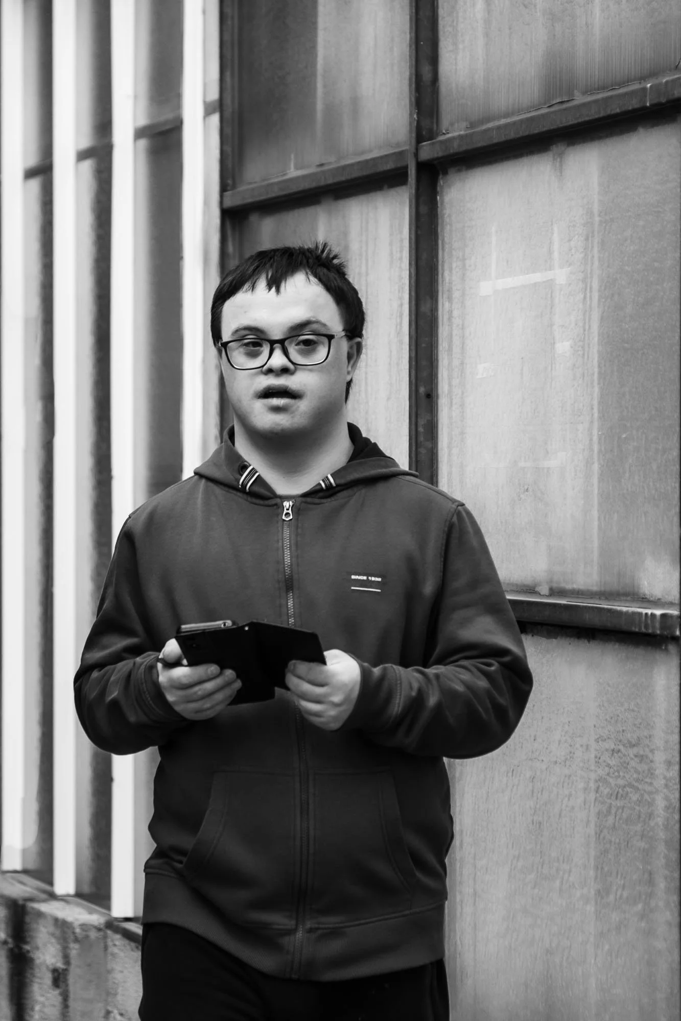 Un giovane uomo con occhiali che tiene un telefono in mano si trova davanti a una parete di metallo, in un'area urbana.