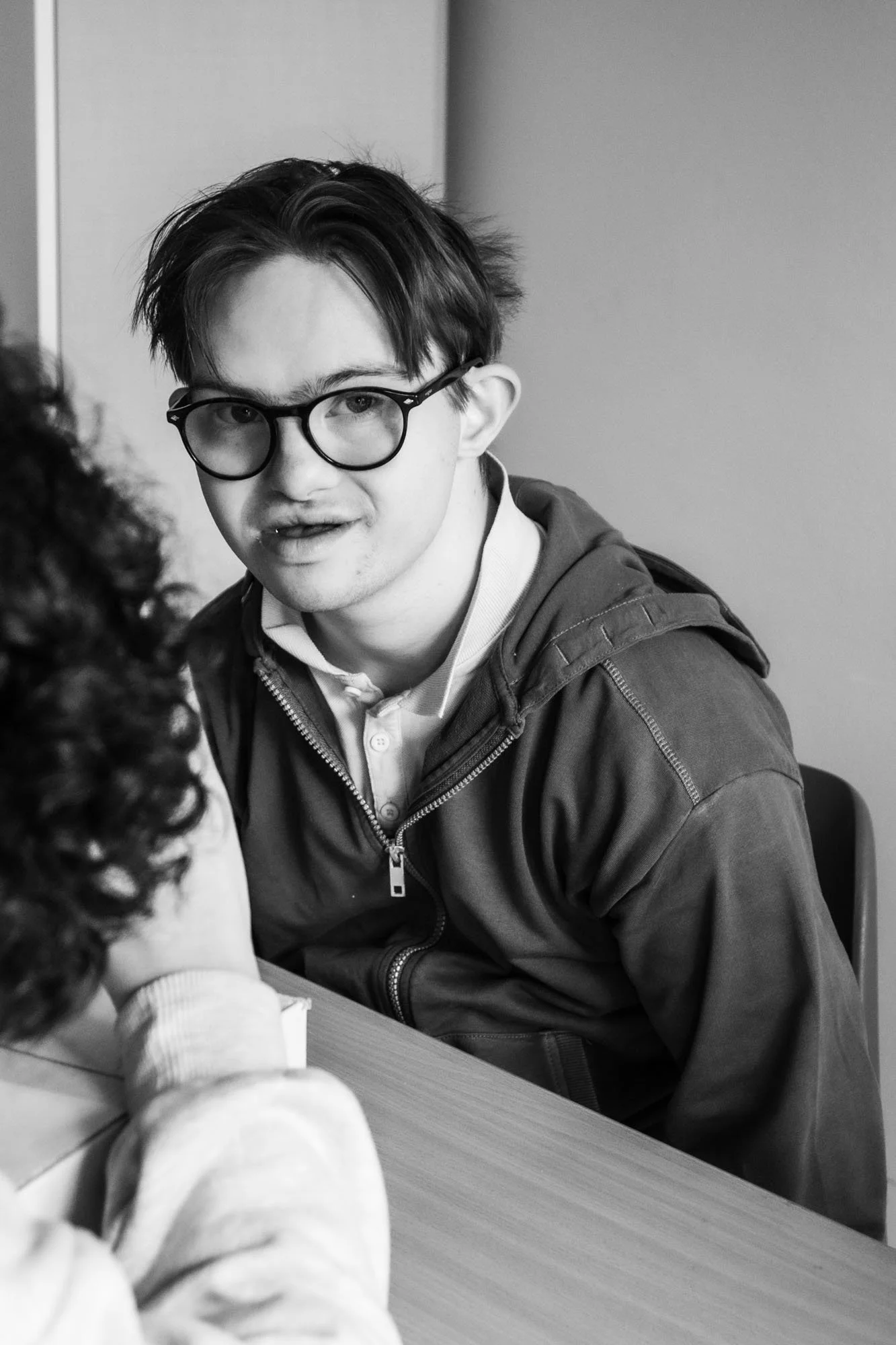 Ragazzo con occhiali, capelli scuri e corti, seduto a un tavolo, che interagisce con una persona con capelli ricci. L'immagine è in bianco e nero.