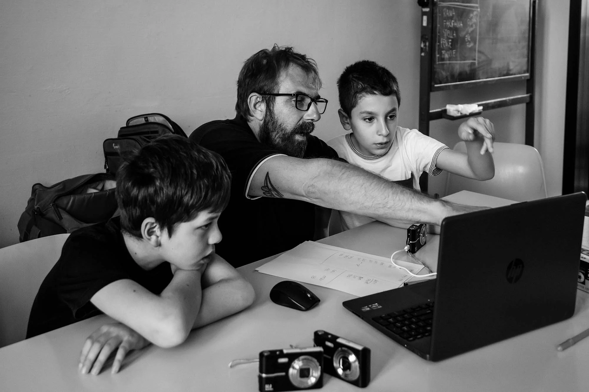 Un adulto e due bambini sono seduti al tavolo, si concentrano su un computer portatile. L'adulto indica qualcosa allo schermo. Sul tavolo ci sono due fotocamere e alcuni fogli di carta.