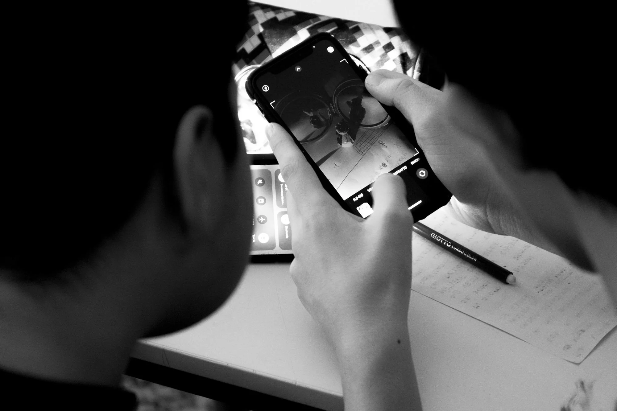 Una persona che scatta una foto con uno smartphone di un disegno su carta, con una penna accanto, in un ambiente di lavoro o studio.