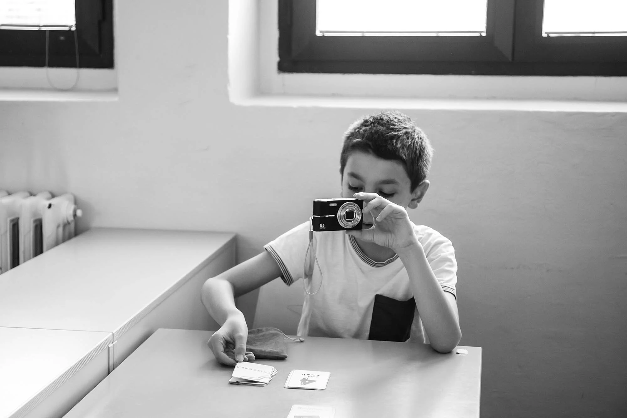 Ragazzo seduto a un tavolo di colori chiari mentre prende una scheda con le istruzioni dell'esercizio e con l'altra mano impugna una macchina fotografica compatta, in un ambiente interno con finestra.