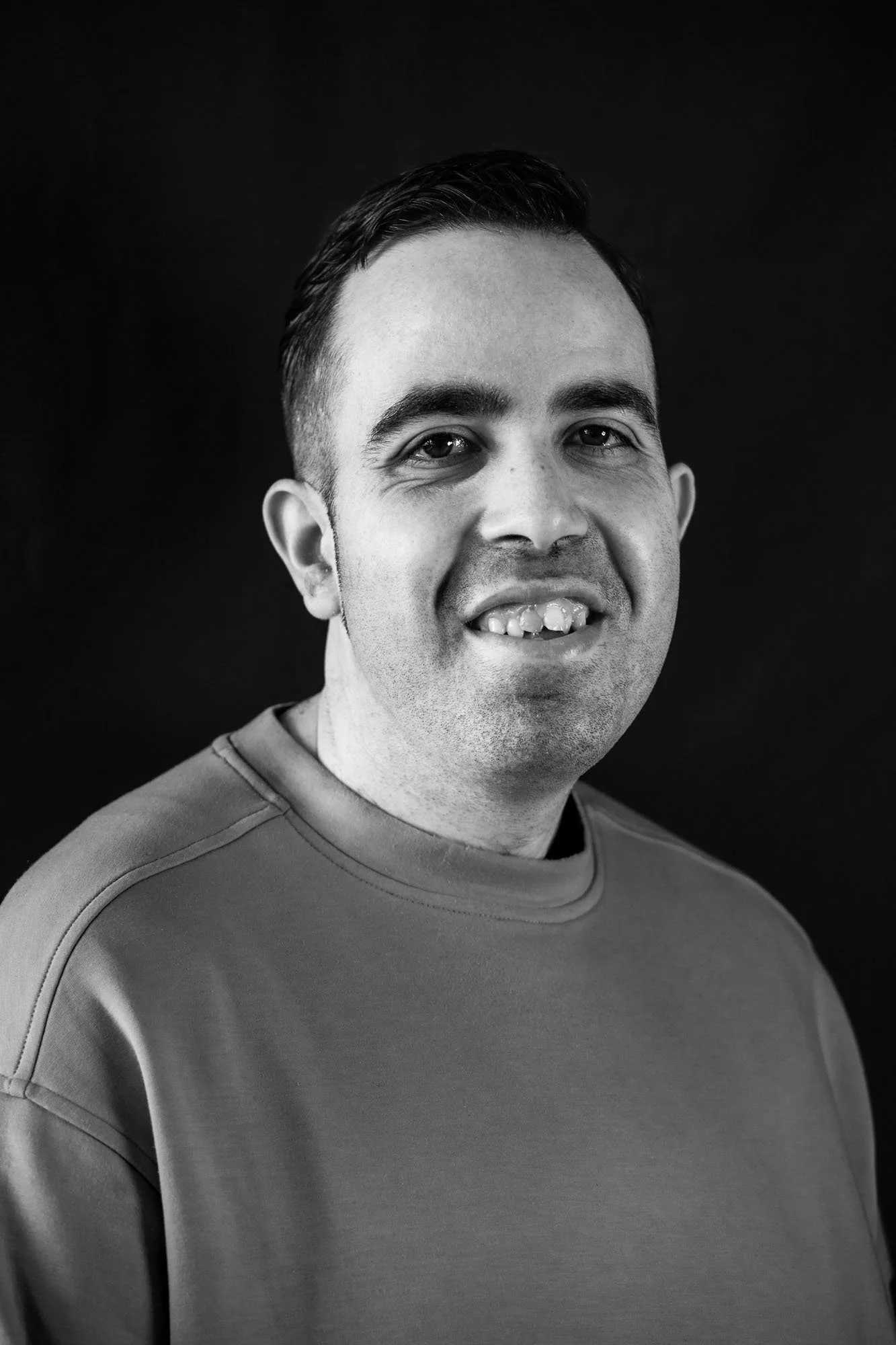 Ritratto di un uomo sorridente con capelli corti e scuri, maglietta collo rotondo, sfondo nero.